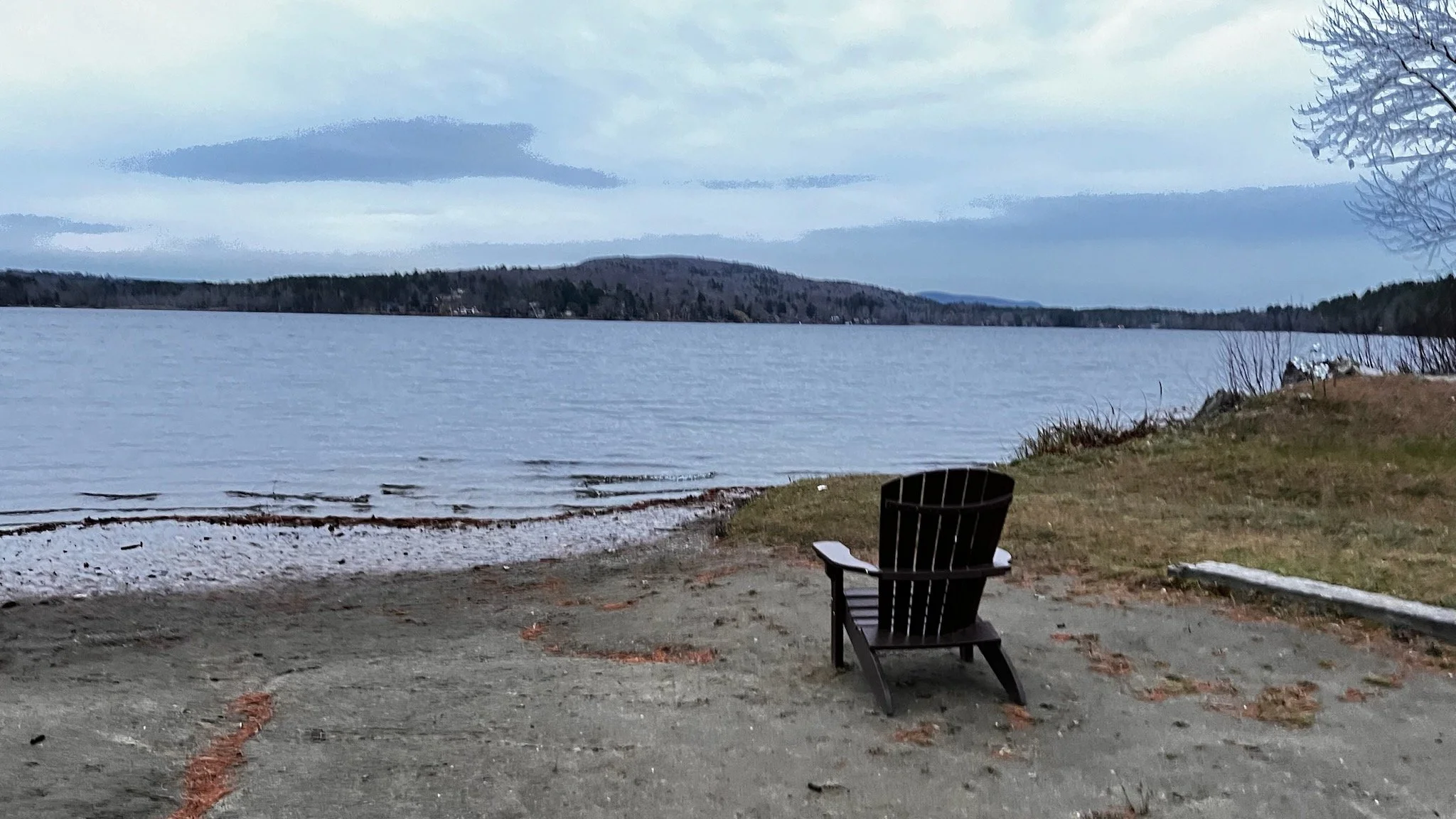 Two women hugging outdoors during sunset, one with brown hair and the other with gray hair wearing a cap and white sweatshirt, with a scenic landscape of trees, mountains, and a lake in the background.
