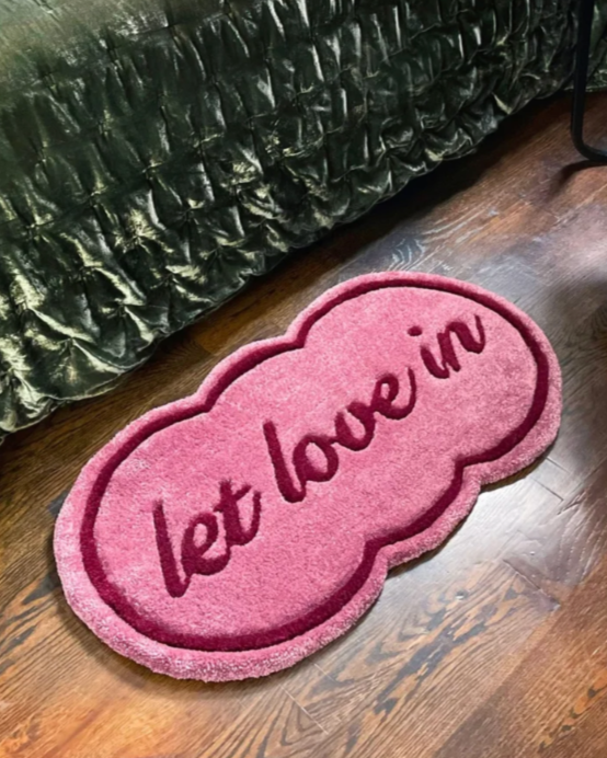 Pink and red rug with the words "let love in" written on it, placed on a wooden floor near a green bedspread.