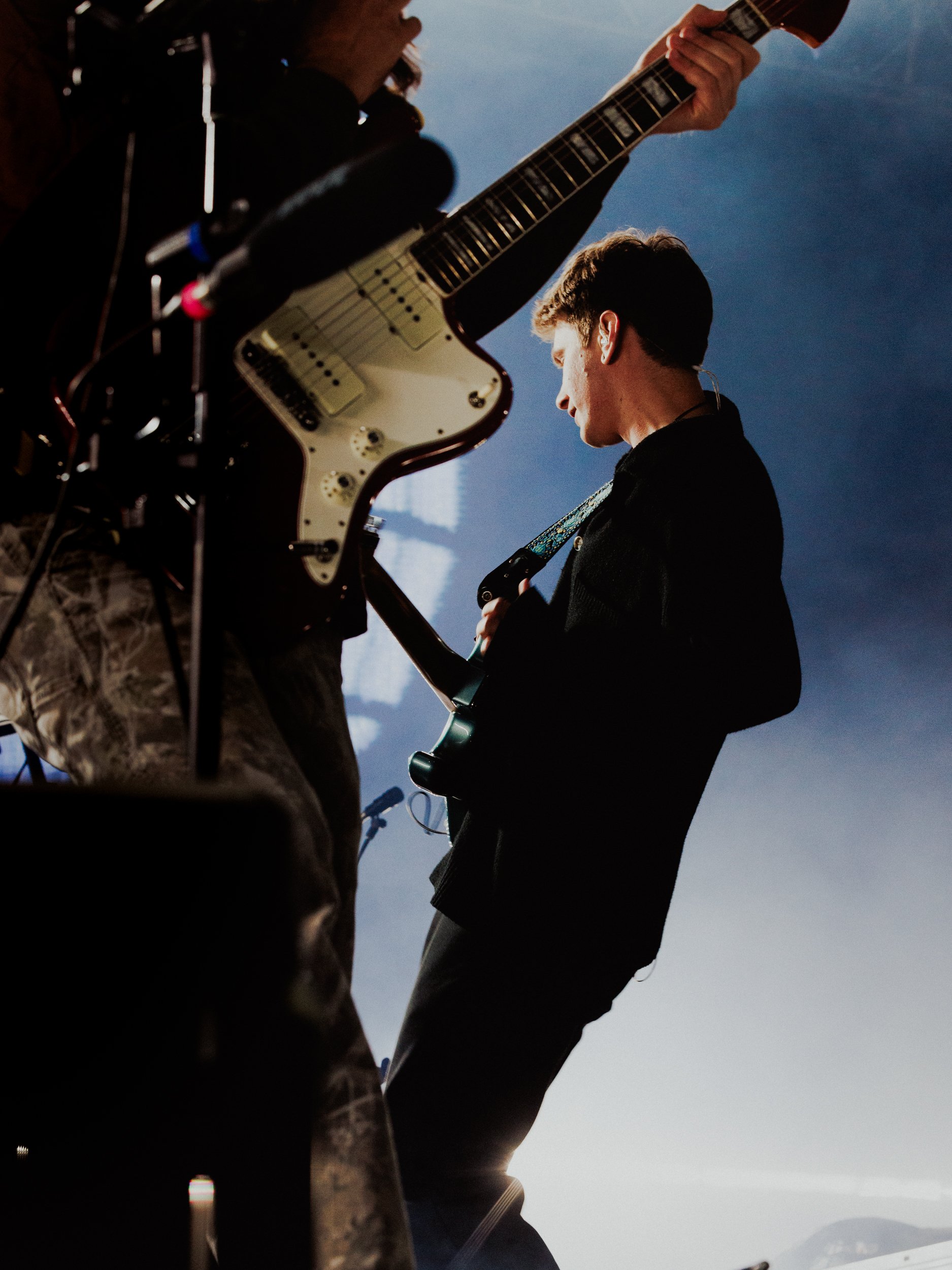 A person playing an electric guitar on stage, viewed from below against a blue sky, with equipment in the foreground.