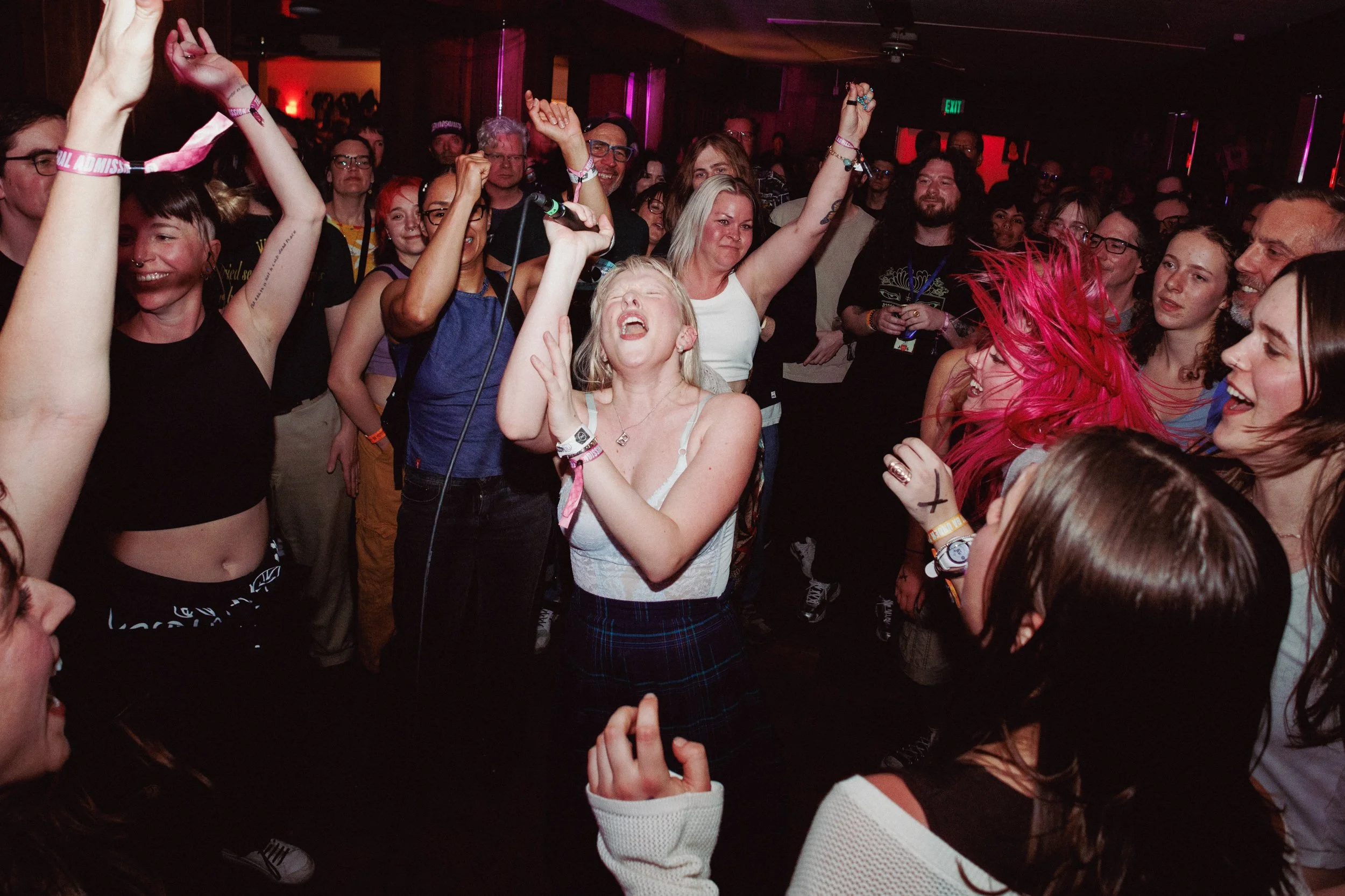 A lively crowd of people dancing and enjoying music in a nightclub or concert venue with dark lighting and colorful neon lights.