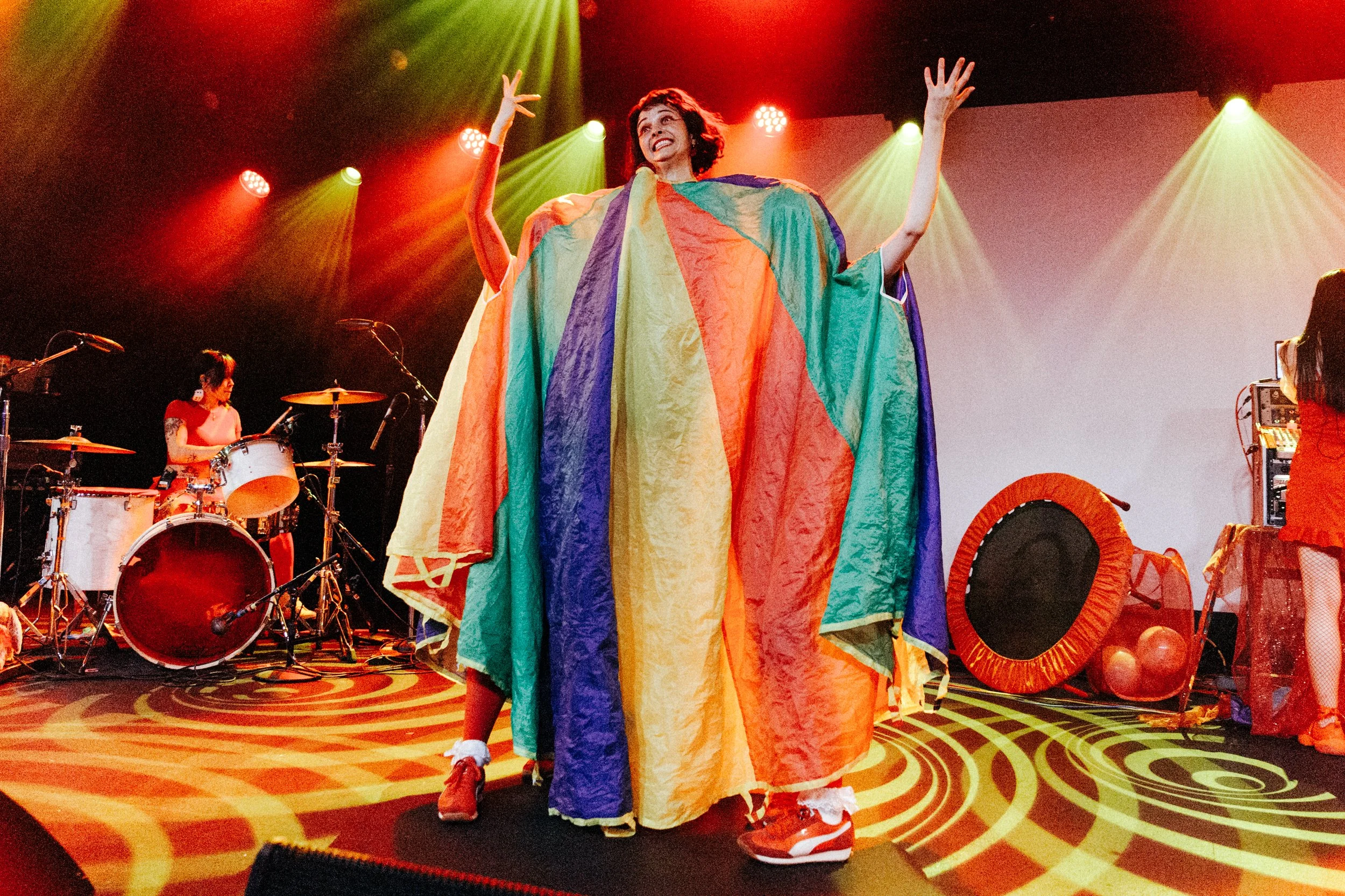 A performer on stage wearing a rainbow-colored cape, smiling and raising her arms, with a drummer and another performer in the background. The stage is lit with red and yellow lights, and the floor has swirling yellow and orange patterns.