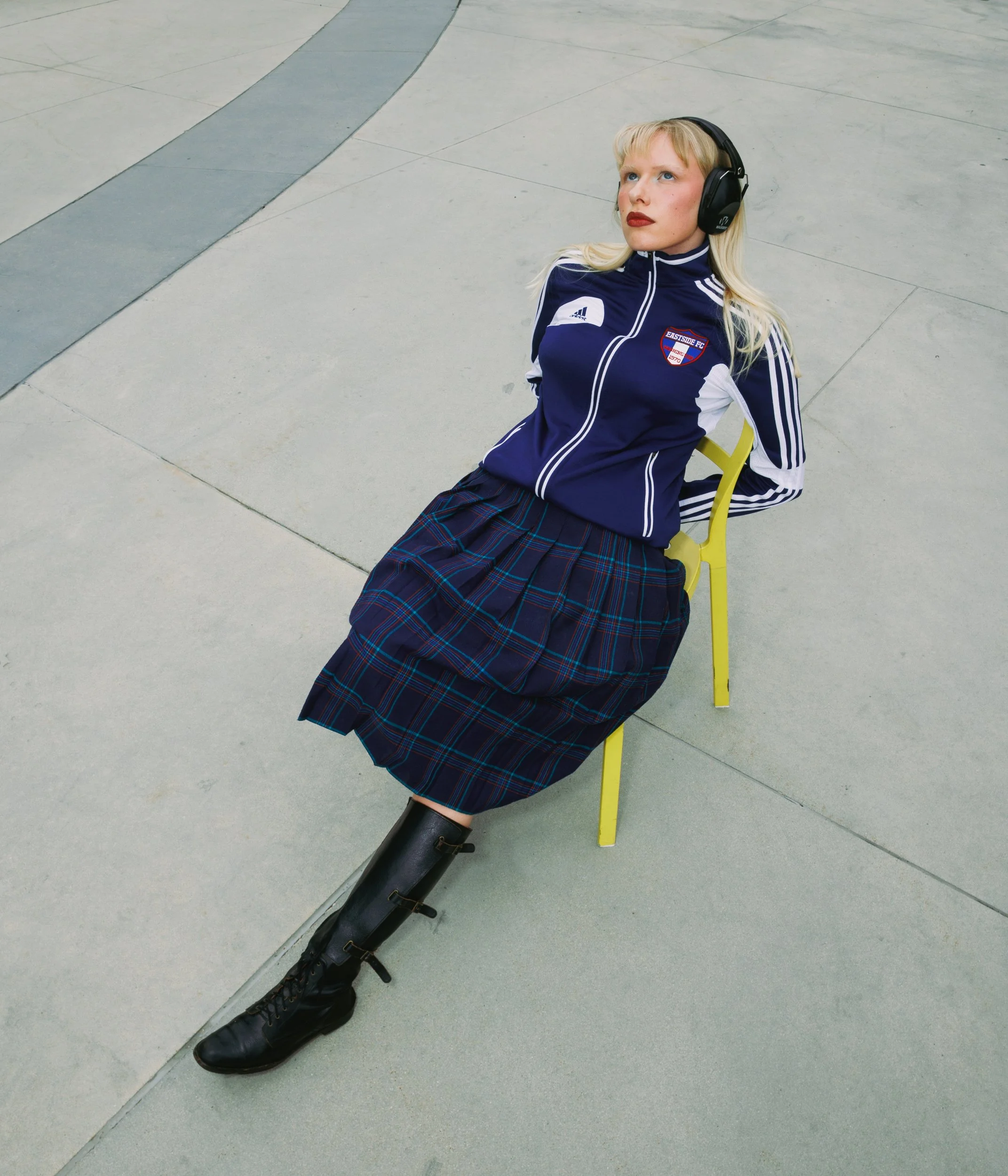 A woman with blonde hair wearing a blue Adidas jacket with football club crest, a tartan skirt, black boots, listening to headphones, seated on a yellow chair on a paved outdoor area.