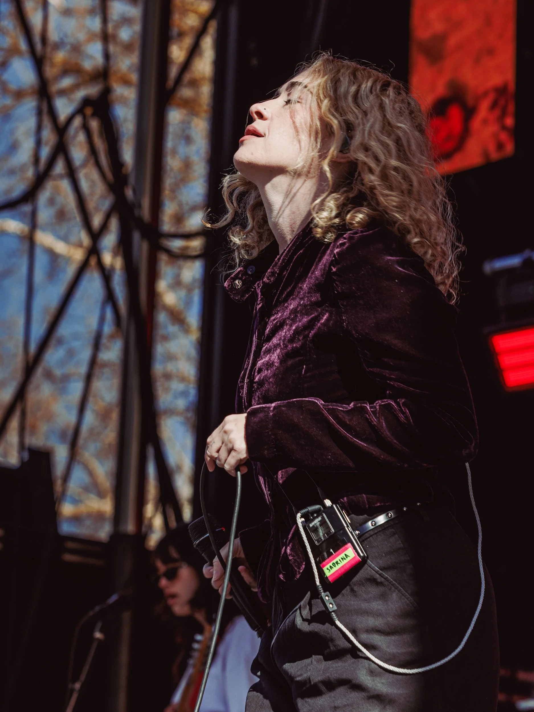 A woman with curly hair singing on stage, wearing a velvet jacket, with a microphone clip attached to her belt, and another person in the background playing guitar.
