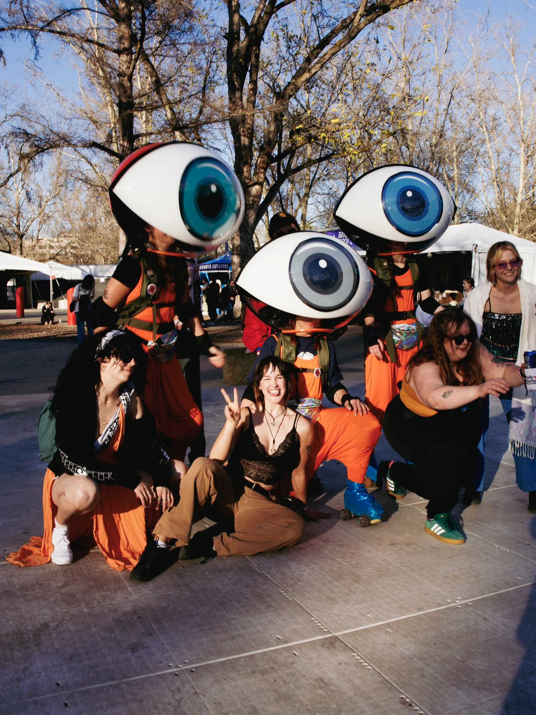 A group of people wearing large eye costumes, some others are smiling and making peace signs, at an outdoor event in a park with leafless trees and tents in the background.