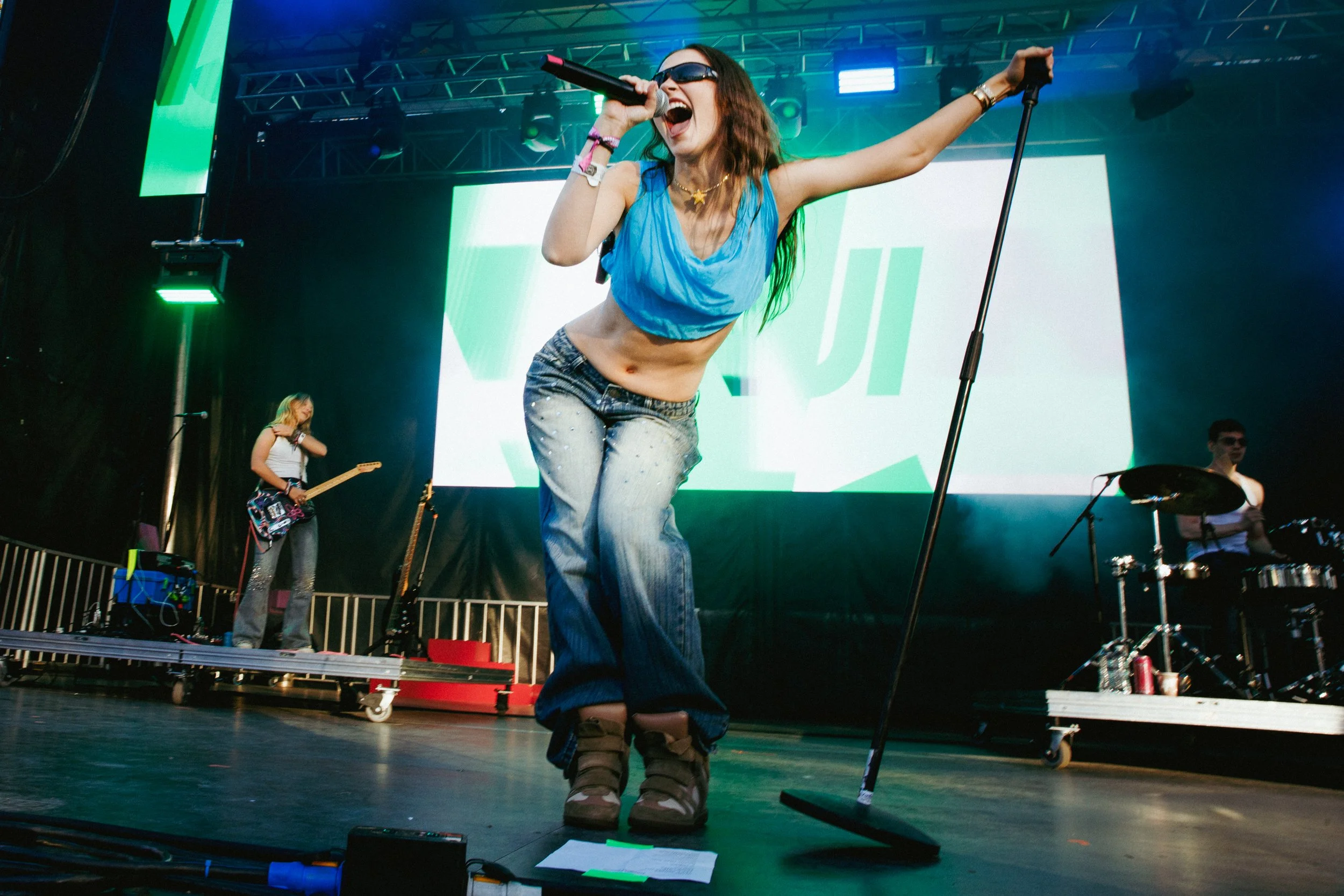 A woman singing passionately on stage with a microphone, wearing sunglasses, a blue top, and jeans. Two other musicians are in the background playing instruments, with stage lights and a large screen behind them.