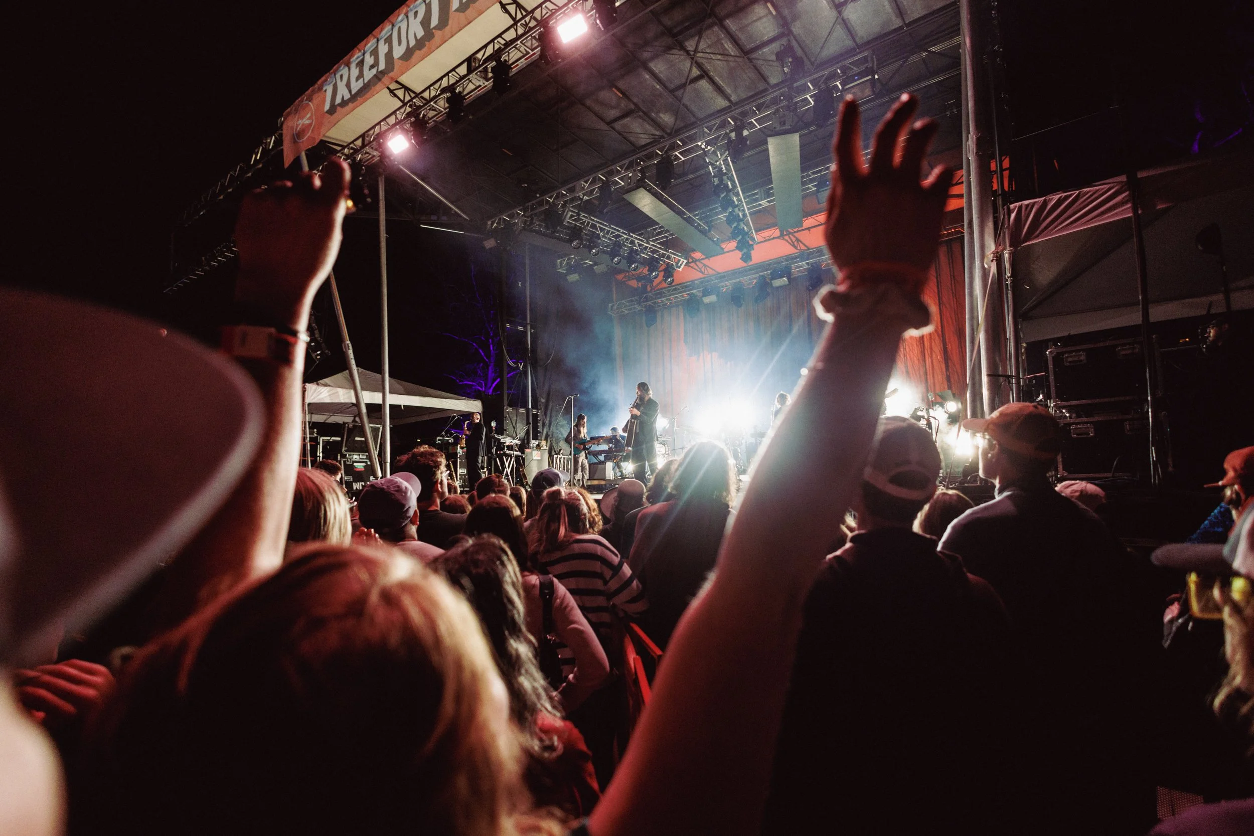 Crowd attending a nighttime outdoor concert with hands raised, watching a band perform on stage illuminated by bright lights.