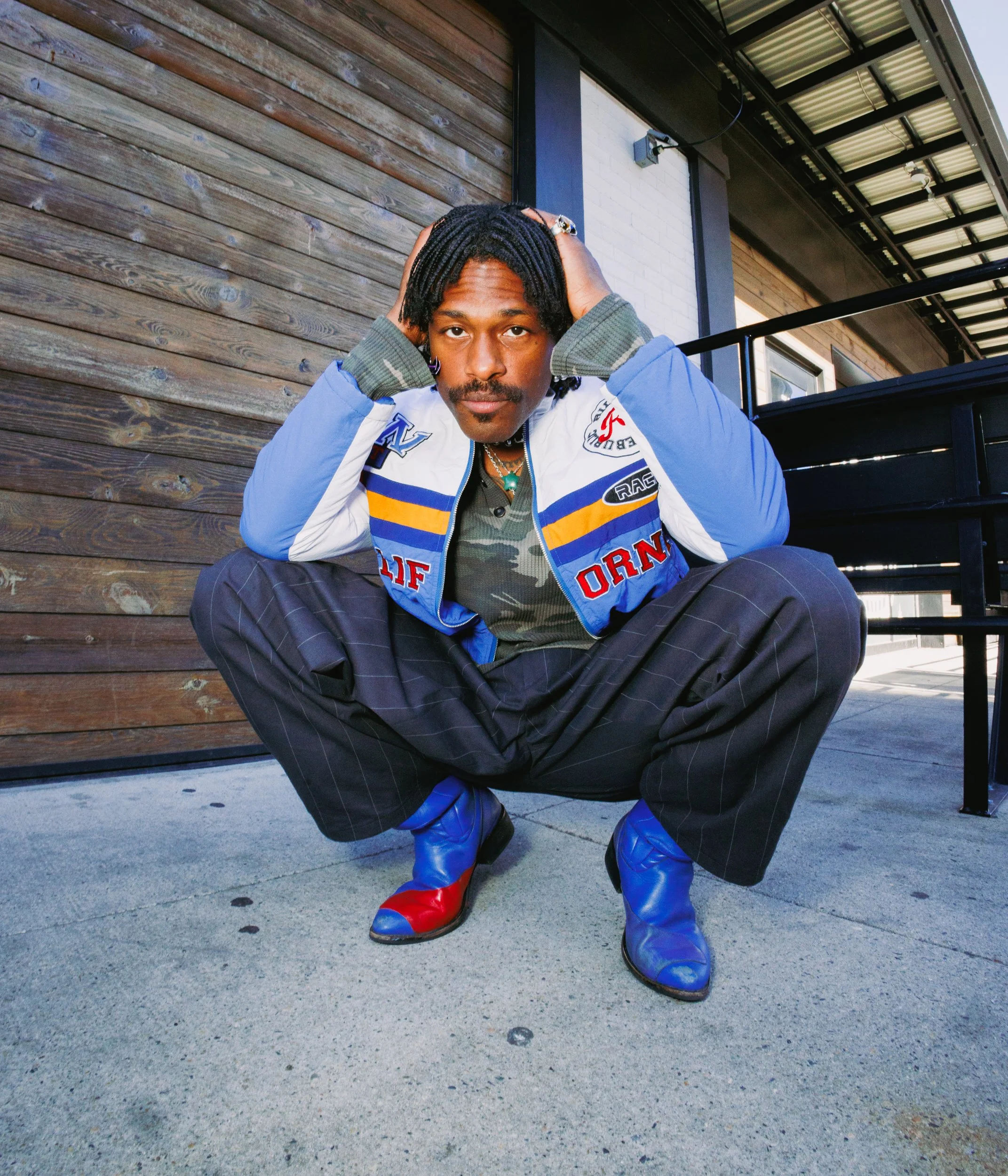 A man squatting outside on a concrete sidewalk, dressed in a colorful patchwork jacket, camouflage shirt, pinstripe pants, and blue and red cowboy boots, with a wooden wall and building in the background.