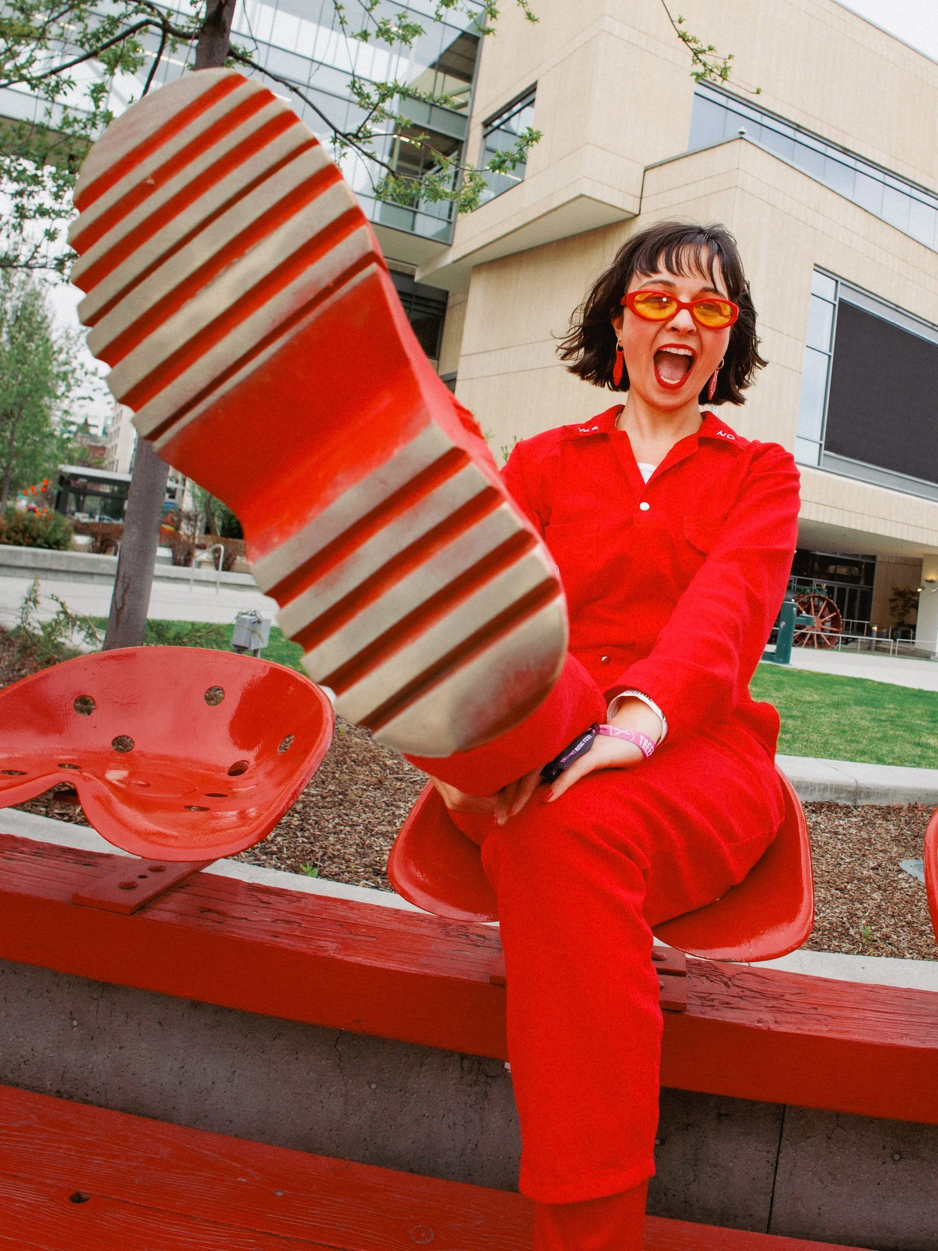 A woman wearing red clothes, sunglasses, and earrings sitting on a red bench outdoors in front of a modern building, laughing and stretching her leg toward the camera with her foot prominently in the foreground.