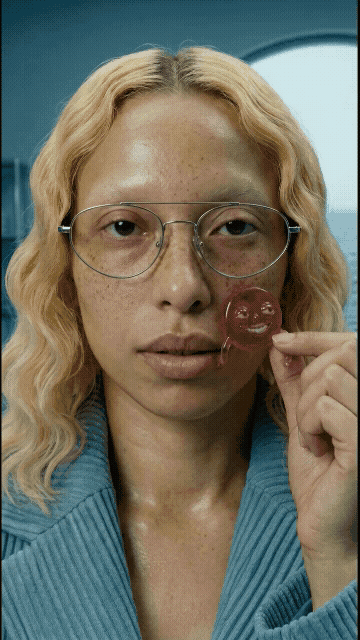 Woman with curly blonde hair and glasses holds a smiley face lollipop close to her face in front of an airplane window.