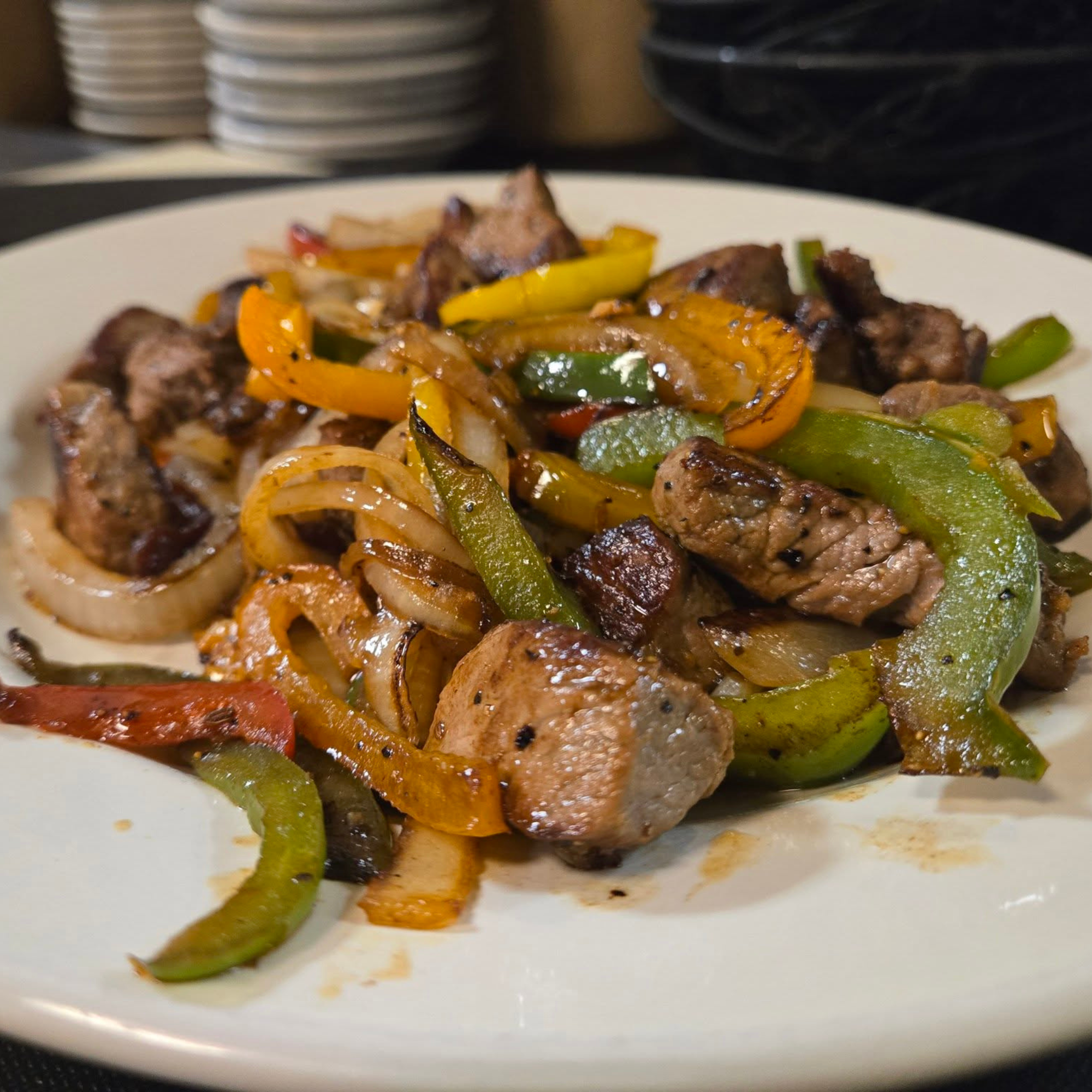 A white plate with grilled beef pieces and mixed sautéed vegetables including green bell peppers, onions, and orange bell peppers.