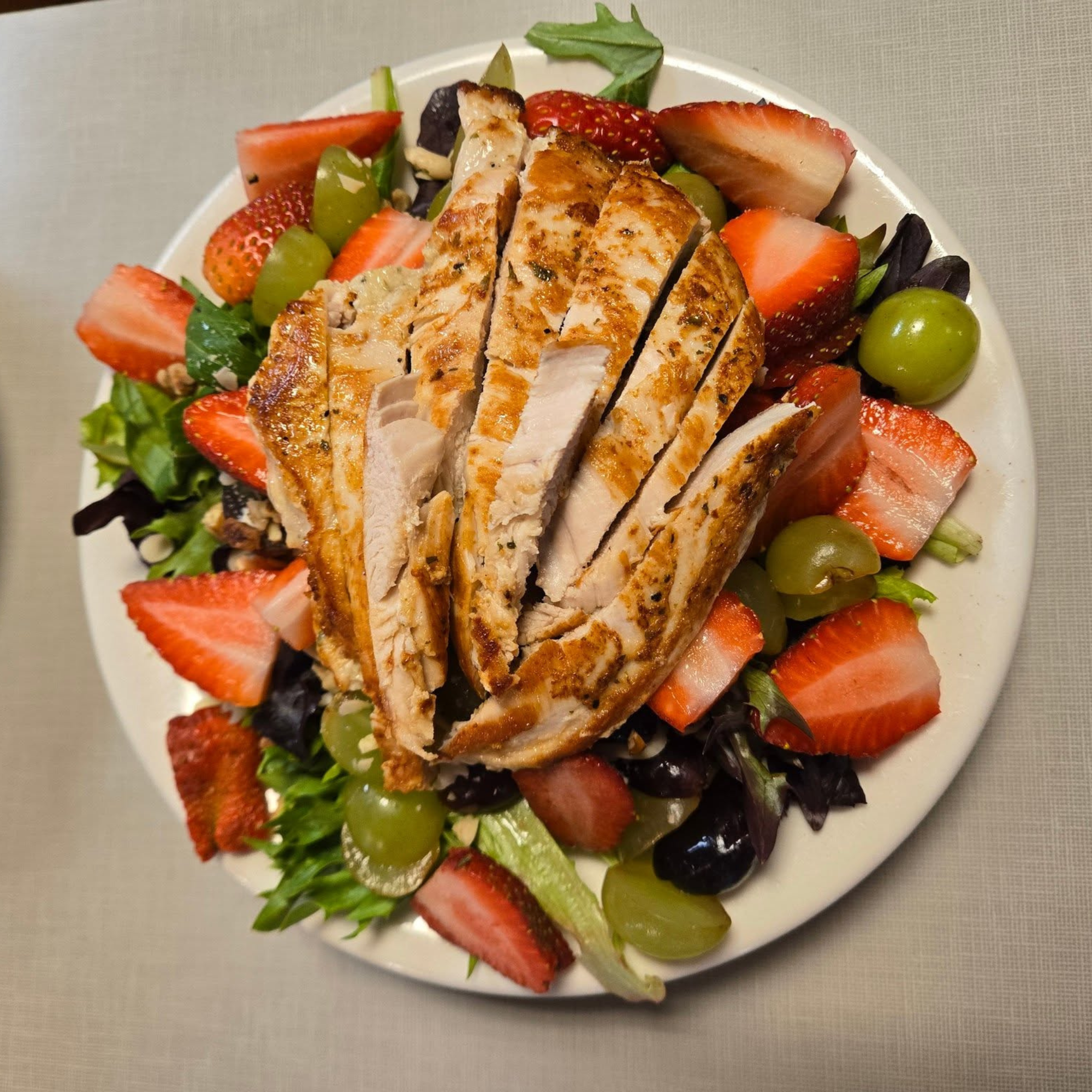 A white plate with grilled chicken breast slices topped with herbs, surrounded by sliced strawberries, green grapes, and mixed greens.