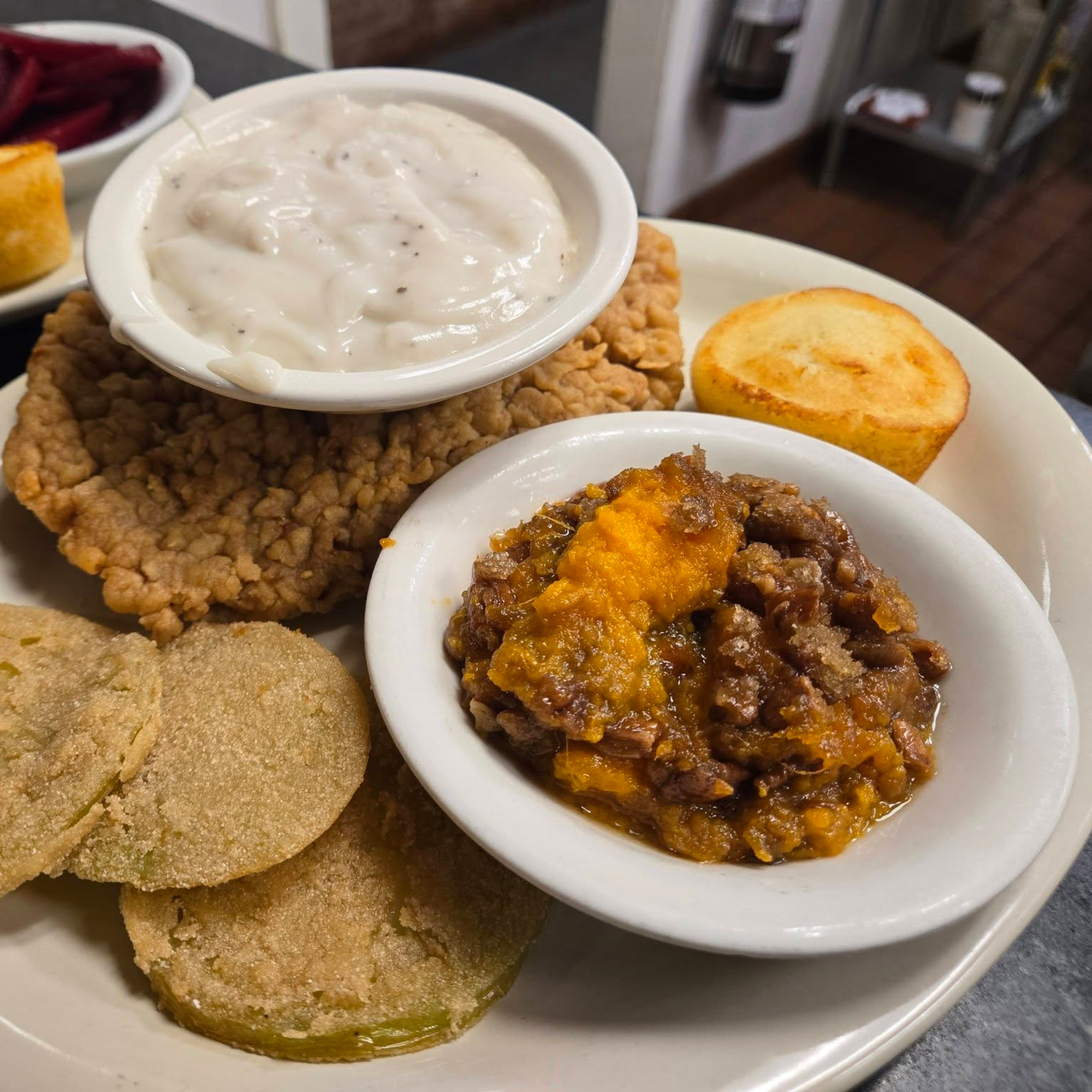 Fried chicken with a side of creamy white gravy in a small bowl, served with baked cornbread and a slice of fried green tomato on a white plate.