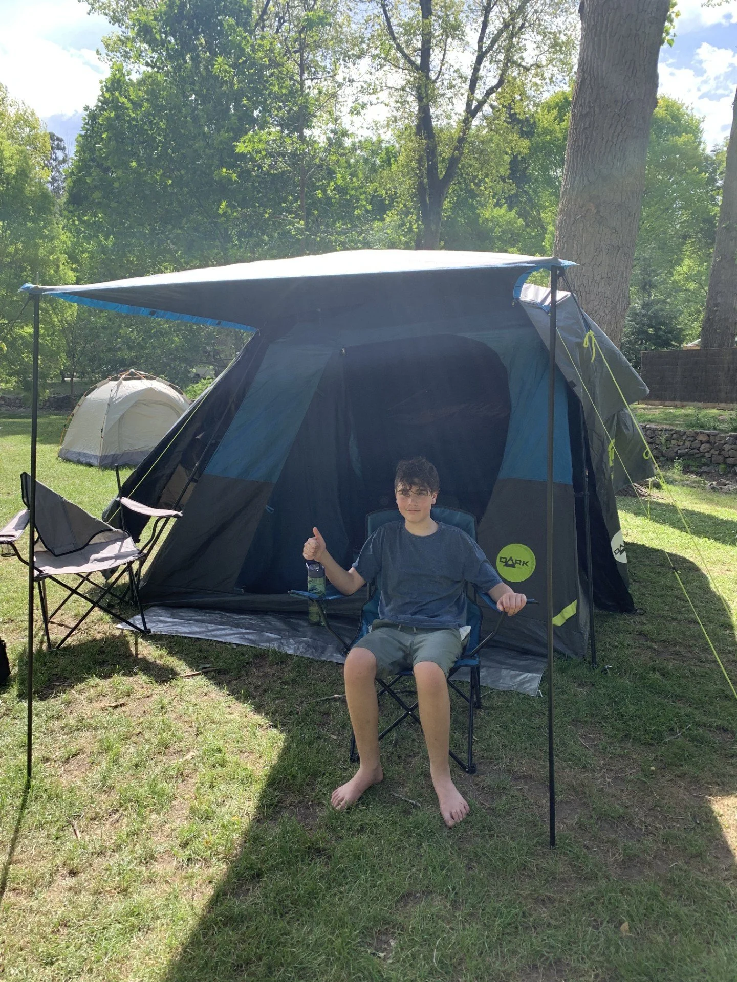 A boy sitting in a camping chair outside a large black and blue tent on a grassy area with trees in the background, holding a water bottle and giving a thumbs-up.