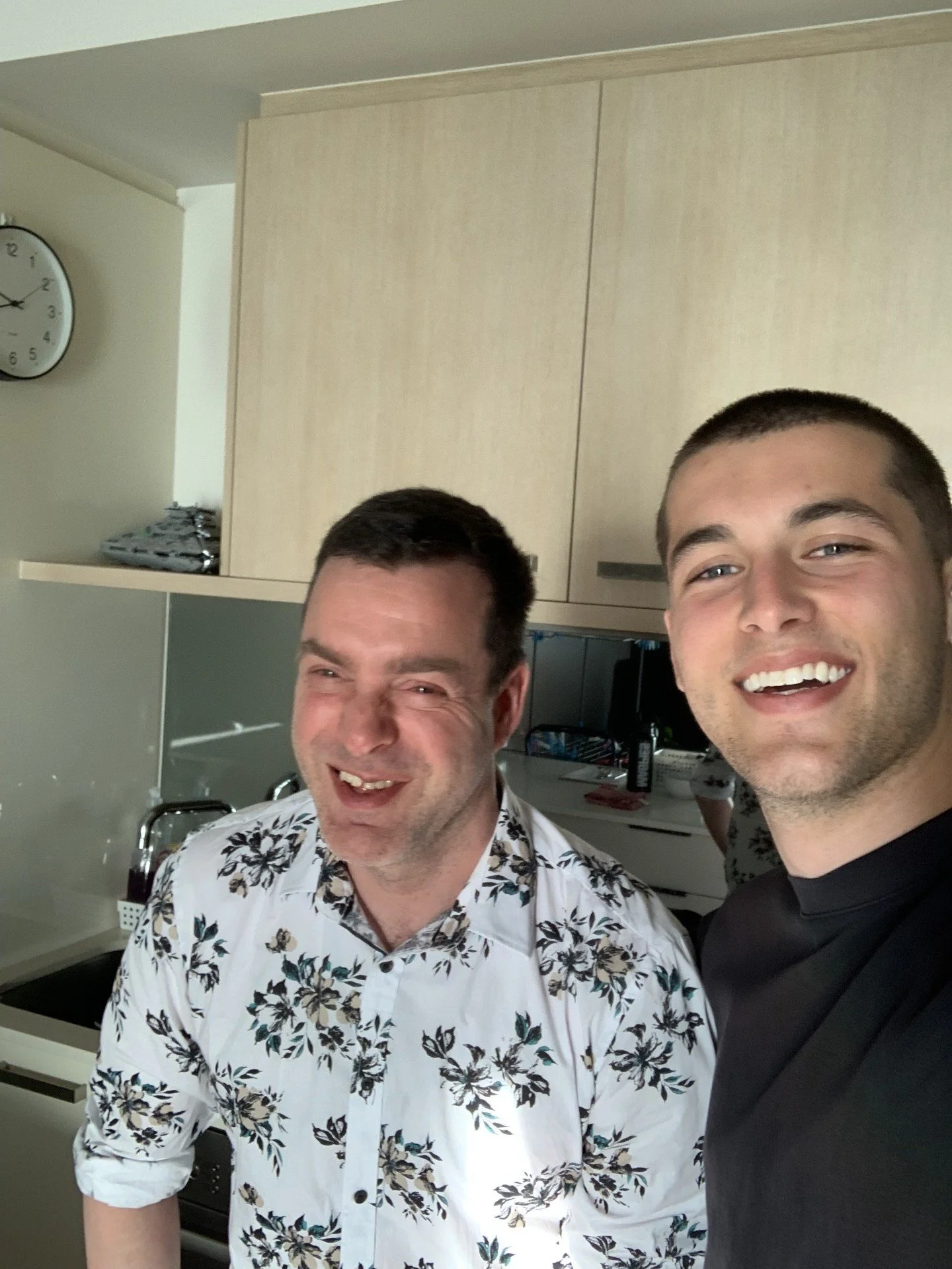 Two men smiling and taking a selfie in a kitchen. One is wearing a white shirt with a black and beige floral pattern, and the other is in a black shirt. A clock and kitchen cabinets are visible in the background.