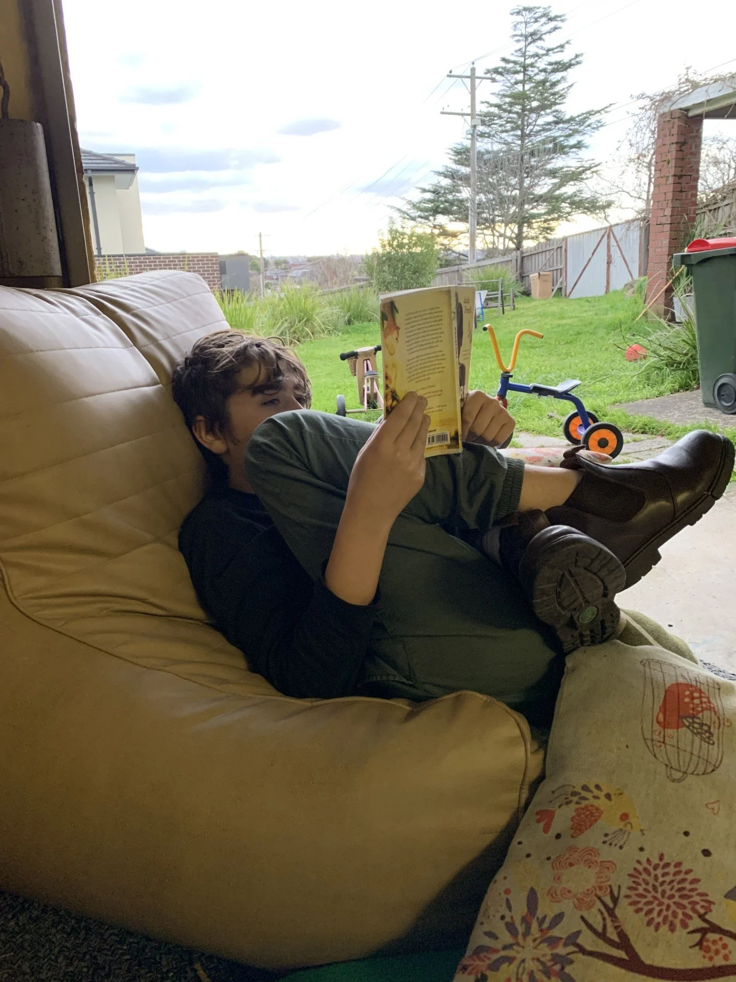 A boy lying on a tan couch, reading a book, with one leg stretched out and the other bent. Outside, there is a grassy backyard with a small child’s tricycle, a green trash bin, and a wooden fence. Overcast sky visible with some clouds.