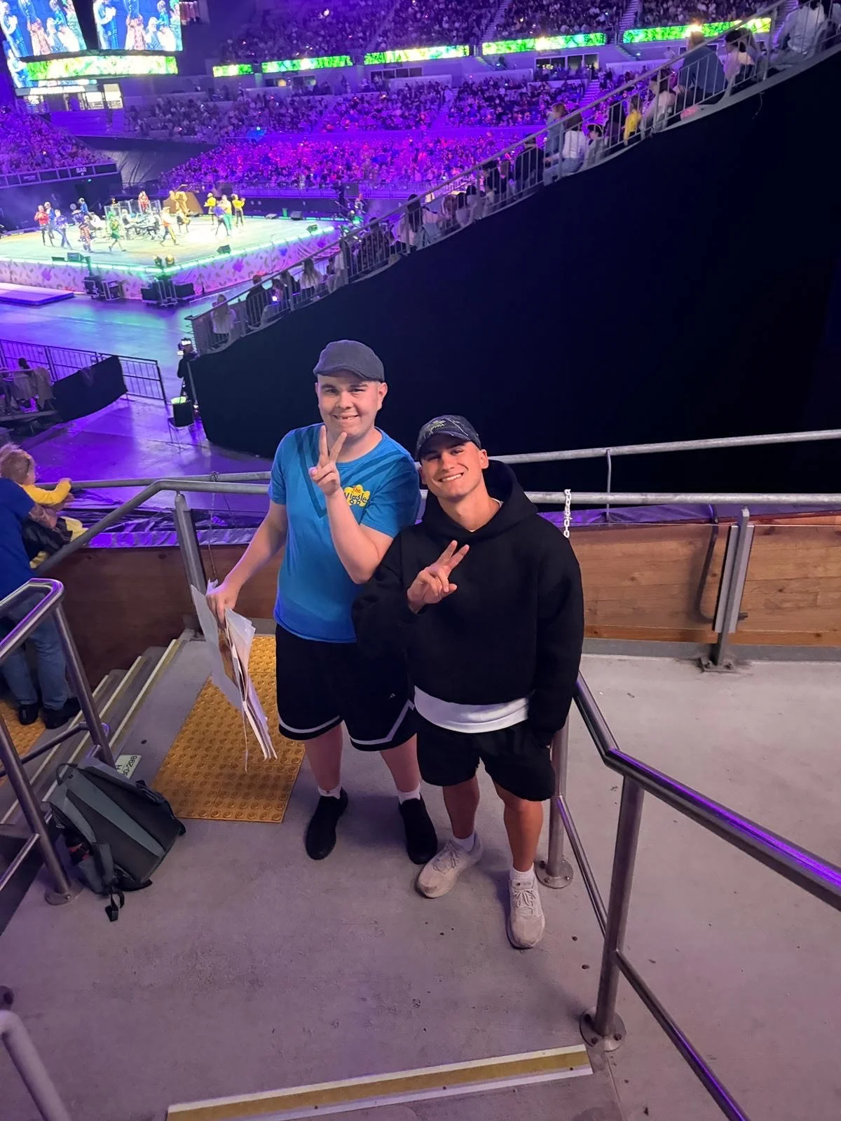 Two young men standing on a staircase at an indoor arena, smiling and making peace signs. The arena has a stage with a dance performance or concert happening, with purple and green lighting, and a large audience seated in the background.