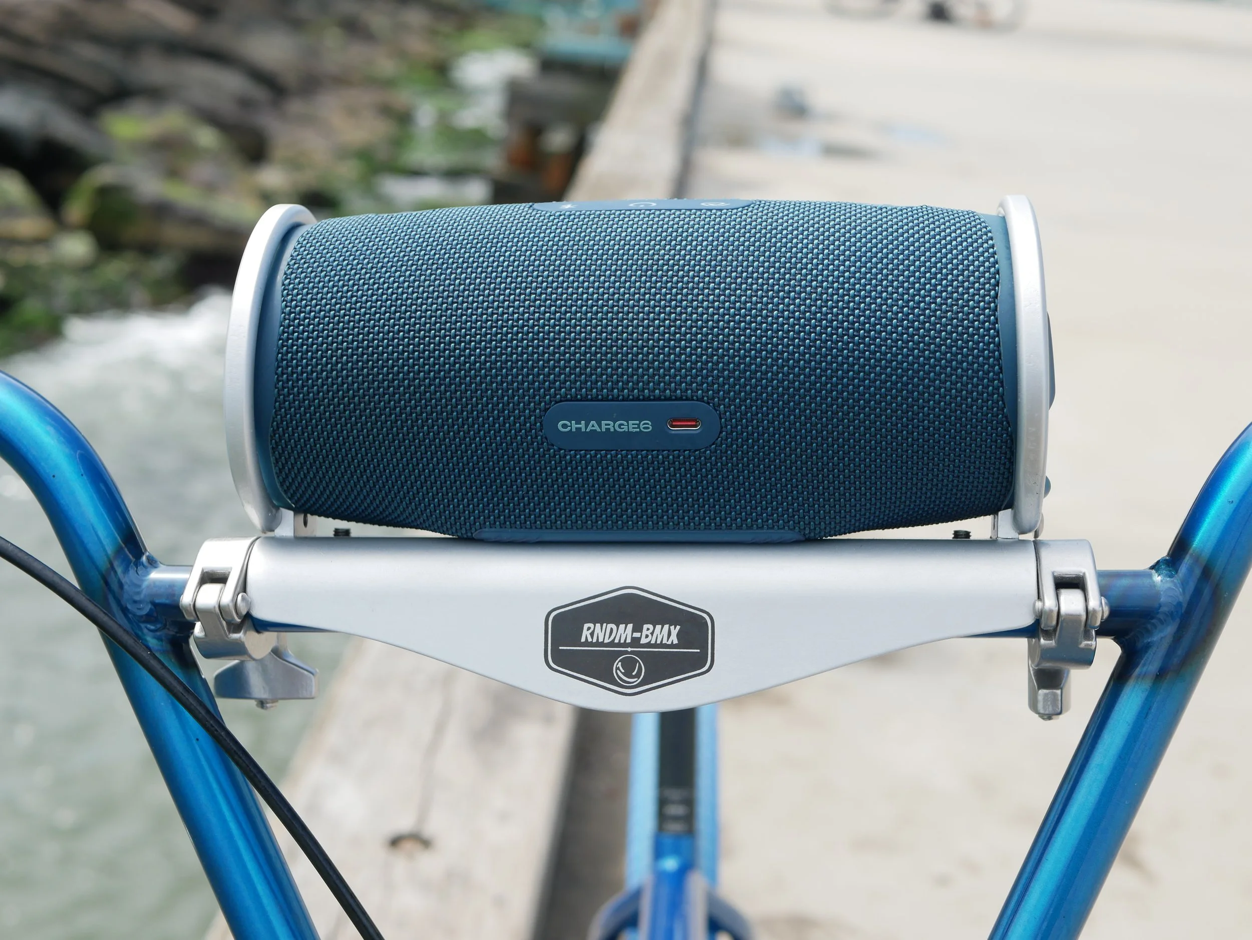 A blue portable Bluetooth speaker with a textured fabric surface, mounted on the handlebars of a bicycle near a waterway.