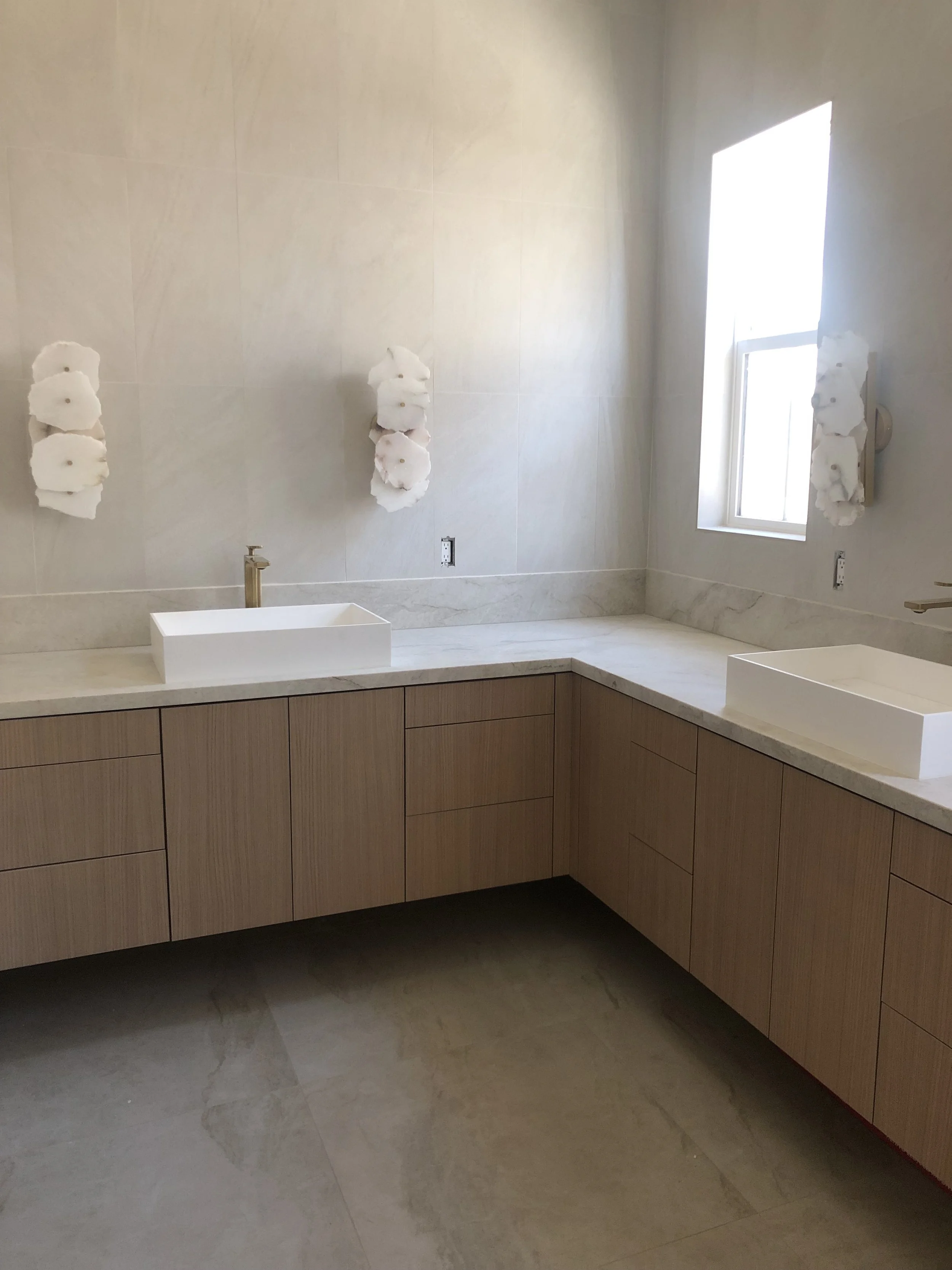 Modern bathroom with light wood cabinets, white countertop, white vessel sinks, and wall-mounted decorative accents natural alabaster stone, carved tiles, taj mahal counter.