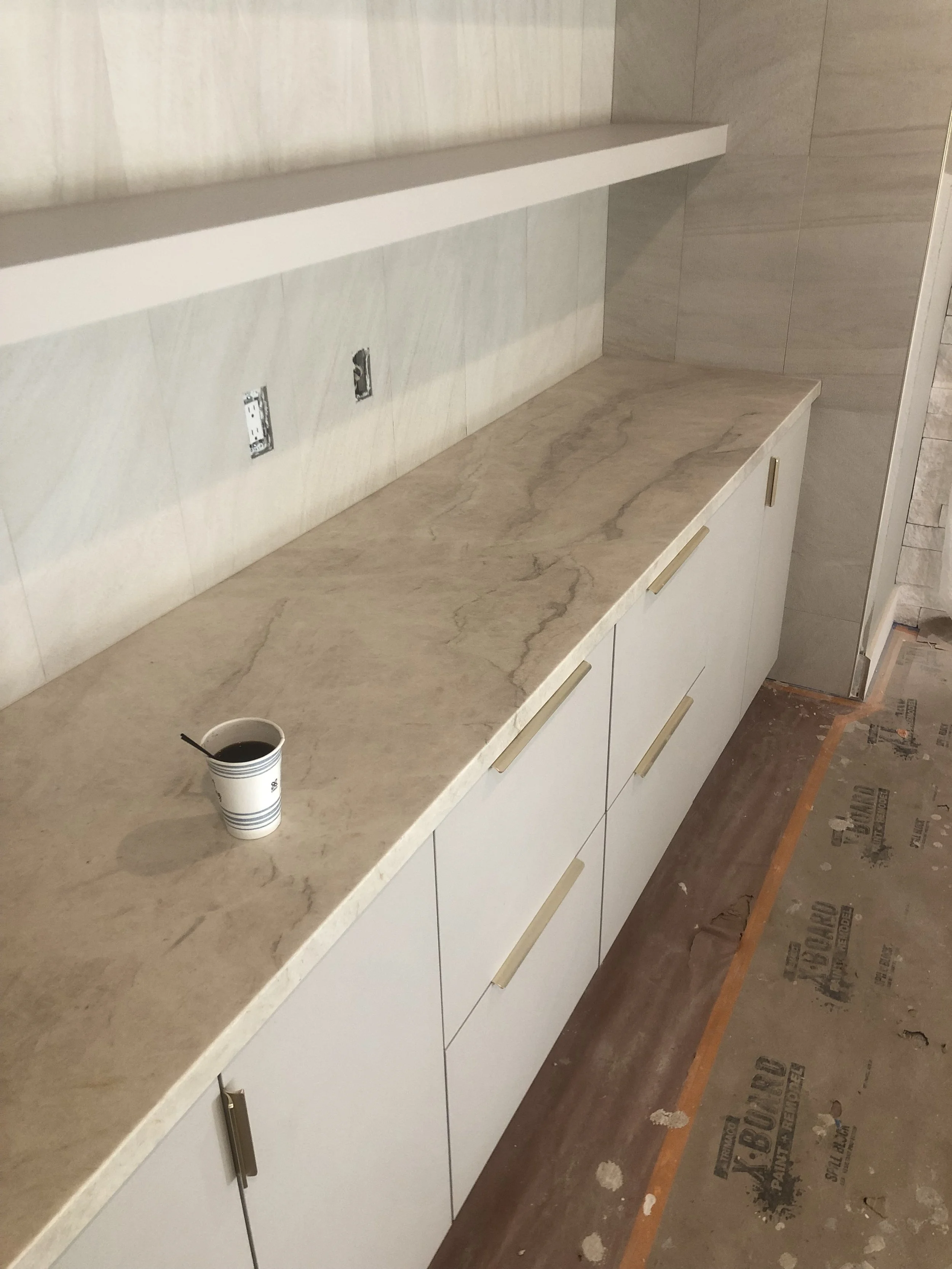 A countertop made of beige taj mahal marble with brown veining, with a cup of coffee. Creme cabinets with gold handles are below