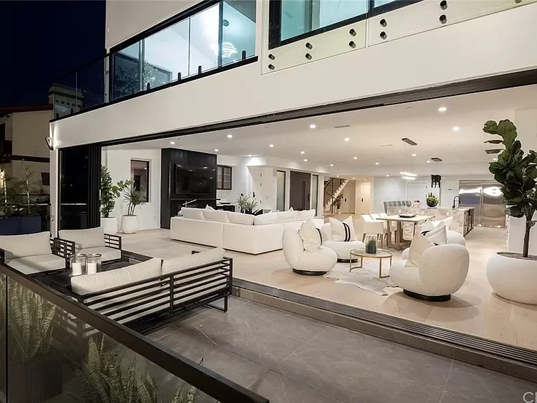 Modern open-plan living room and dining area with white furniture, large potted plants, and a staircase in the background, seen through a large glass window. Newport beach luxury beach front home, custom build.