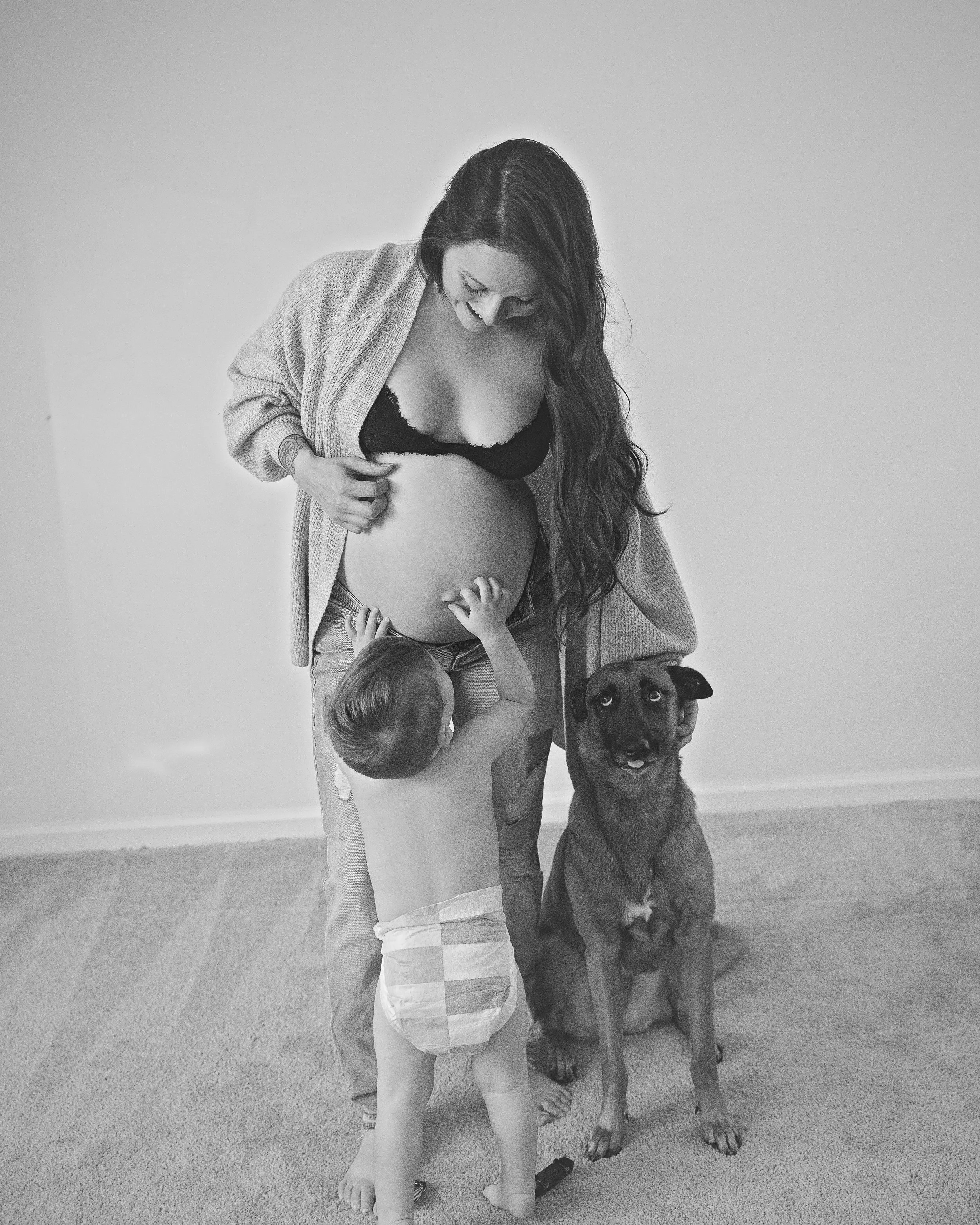 A woman in a gray cardigan and crop top, standing on carpeted floor, interacts with two young children and a large brown dog in a plain room.