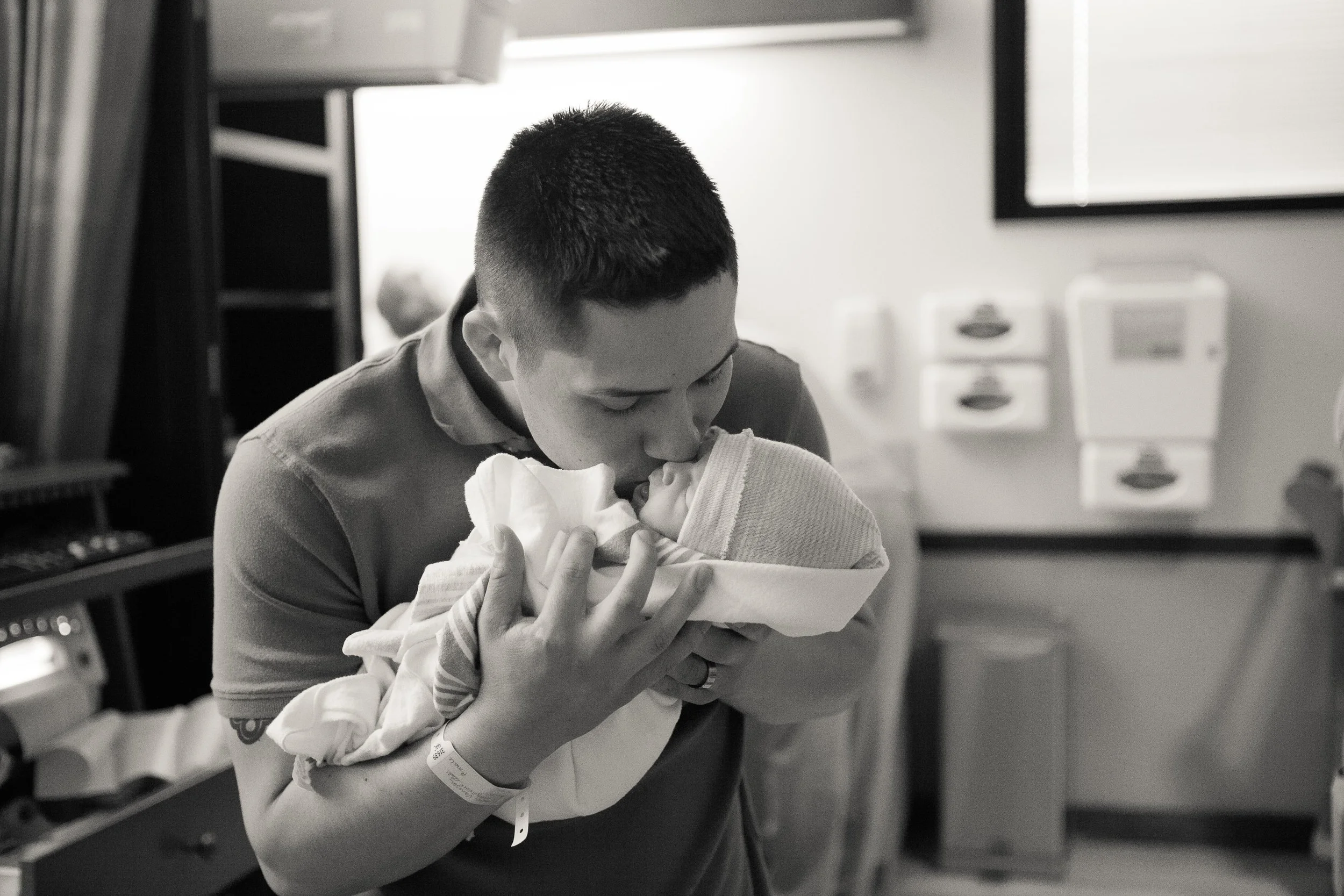 A nurse in a hospital holding a newborn baby close to her face, in a clinical setting with medical equipment in the background.