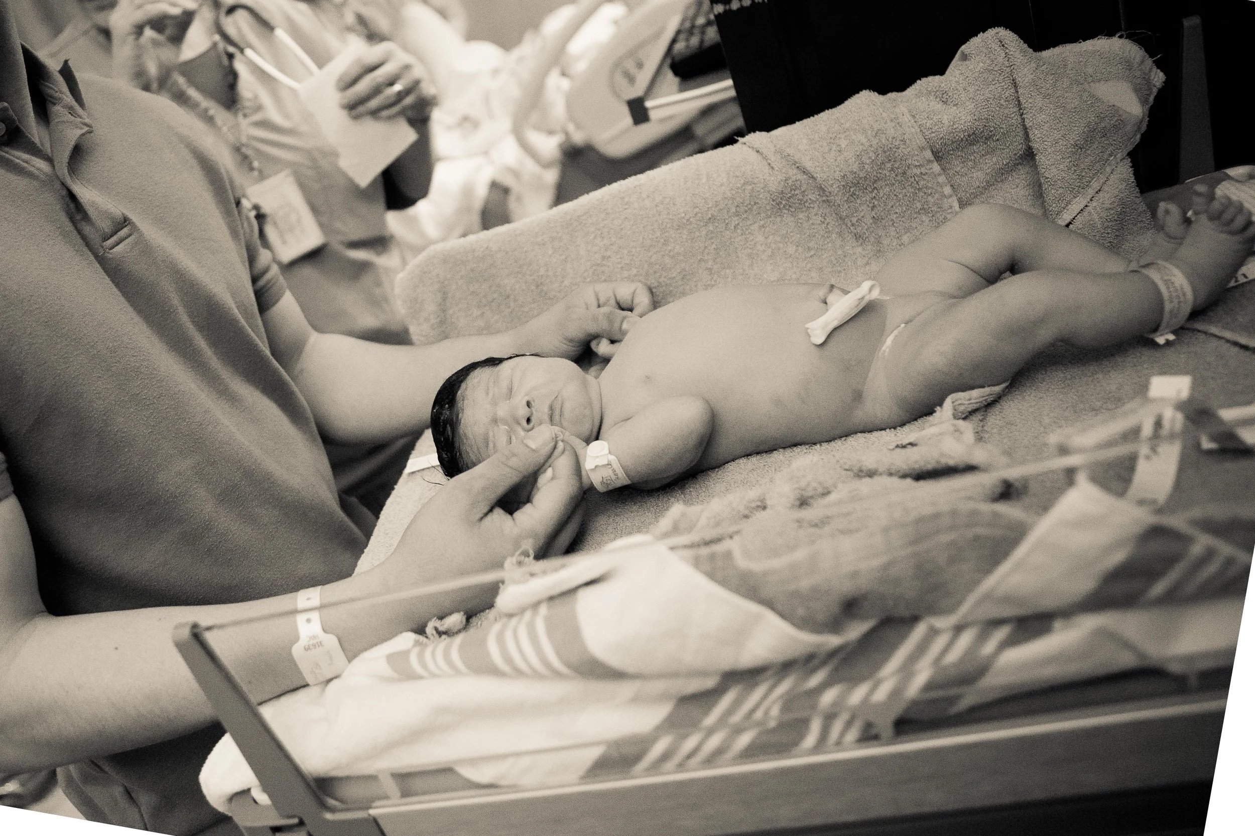 A newborn baby lying on a hospital bed, being held by a person, with medical staff in the background in a hospital setting.