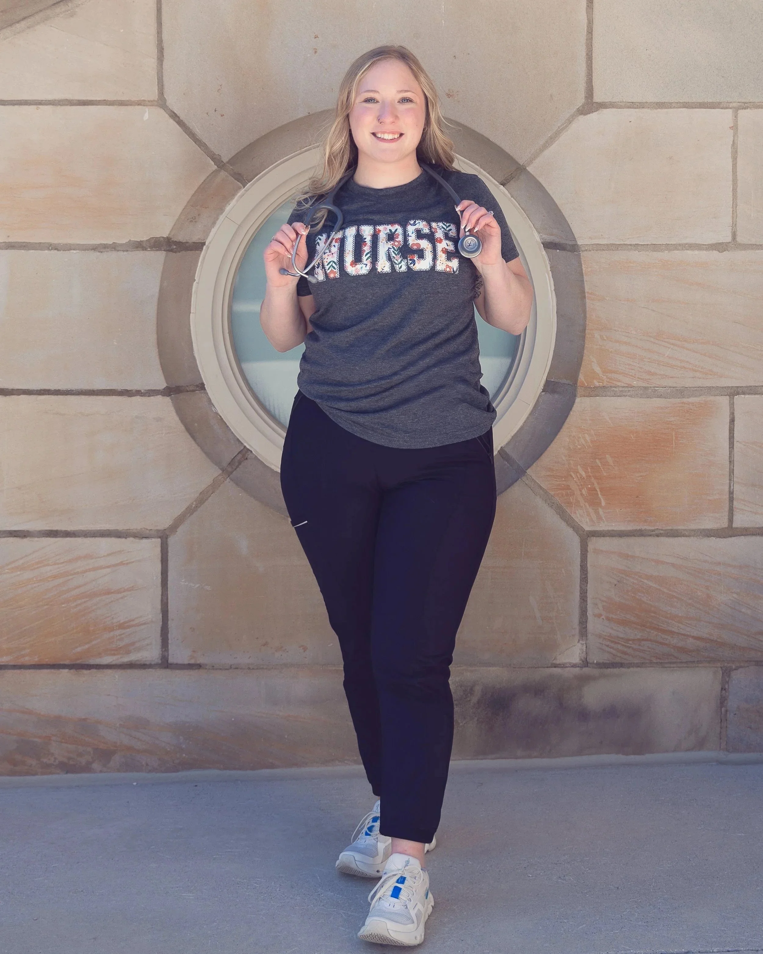 A young woman standing outdoors in front of a stone wall, wearing a dark gray T-shirt with the word 'MUSE' on it, black pants, and white sneakers, holding a stethoscope around her neck, smiling at the camera.