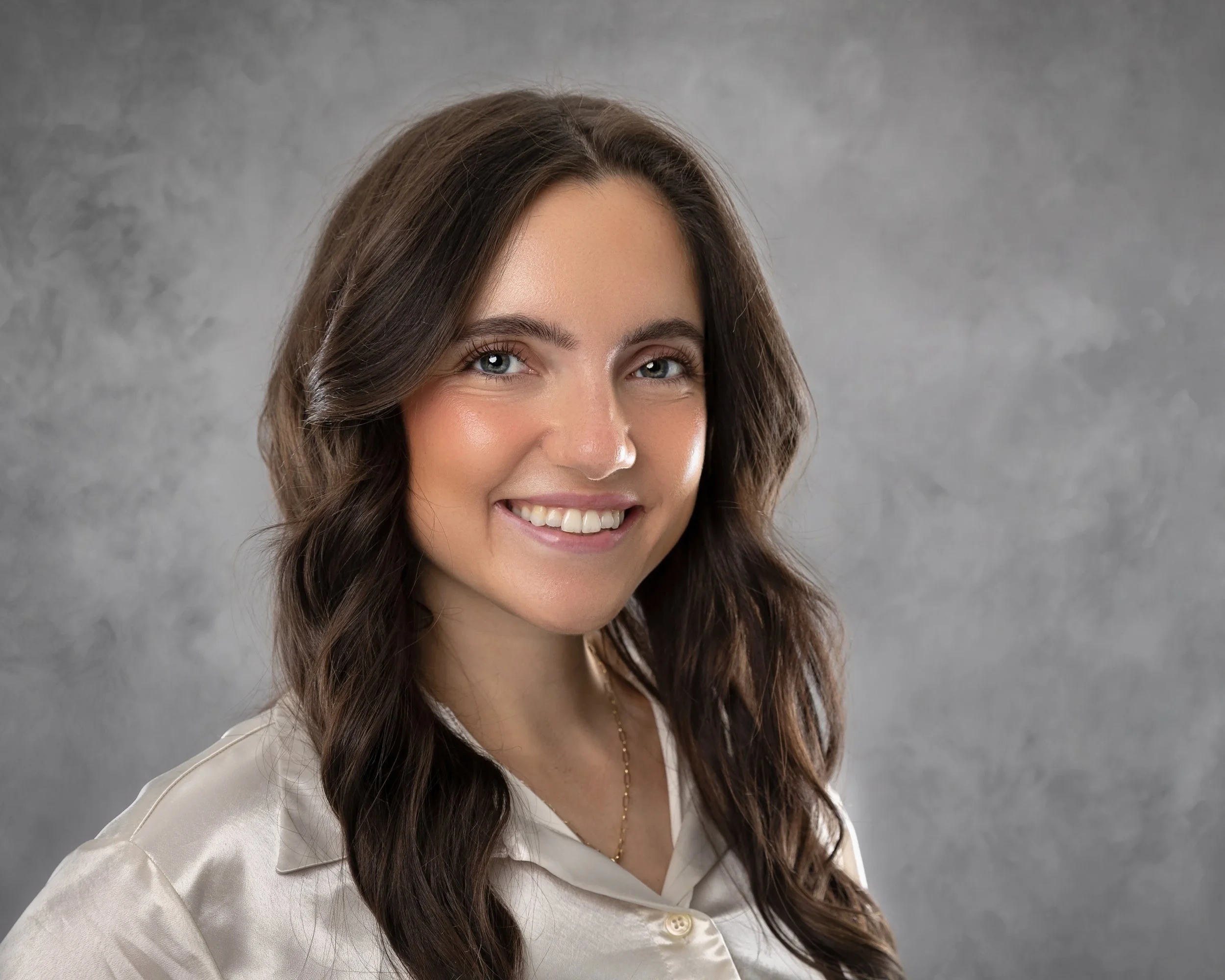 Close-up of a smiling woman with long, wavy brown hair, wearing a beige blazer and white blouse, standing against a gray textured background.