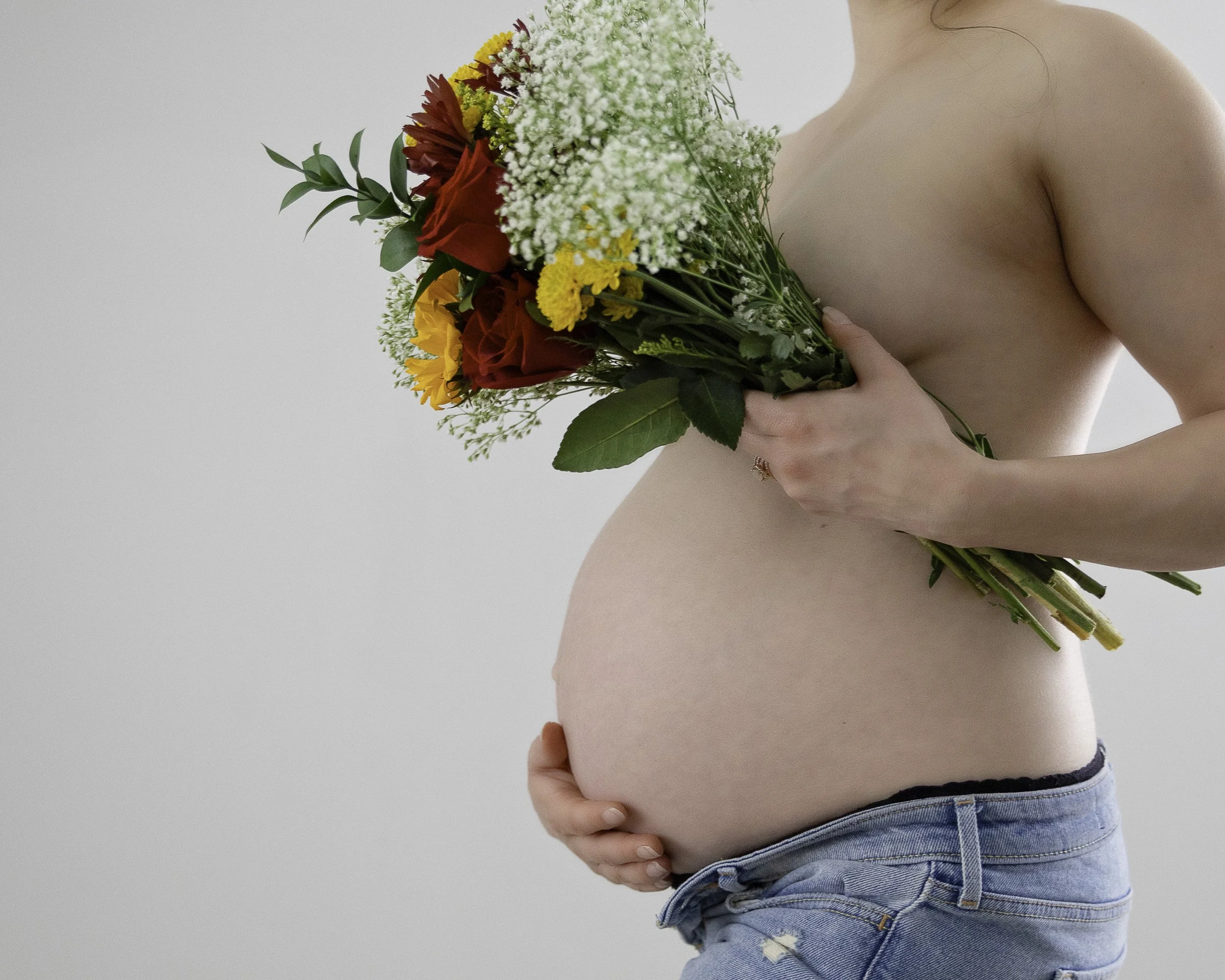A pregnant woman holding a colorful bouquet of flowers in front of her chest, with her other hand resting on her baby bump, wearing jeans and showing her bare shoulder against a plain light background.