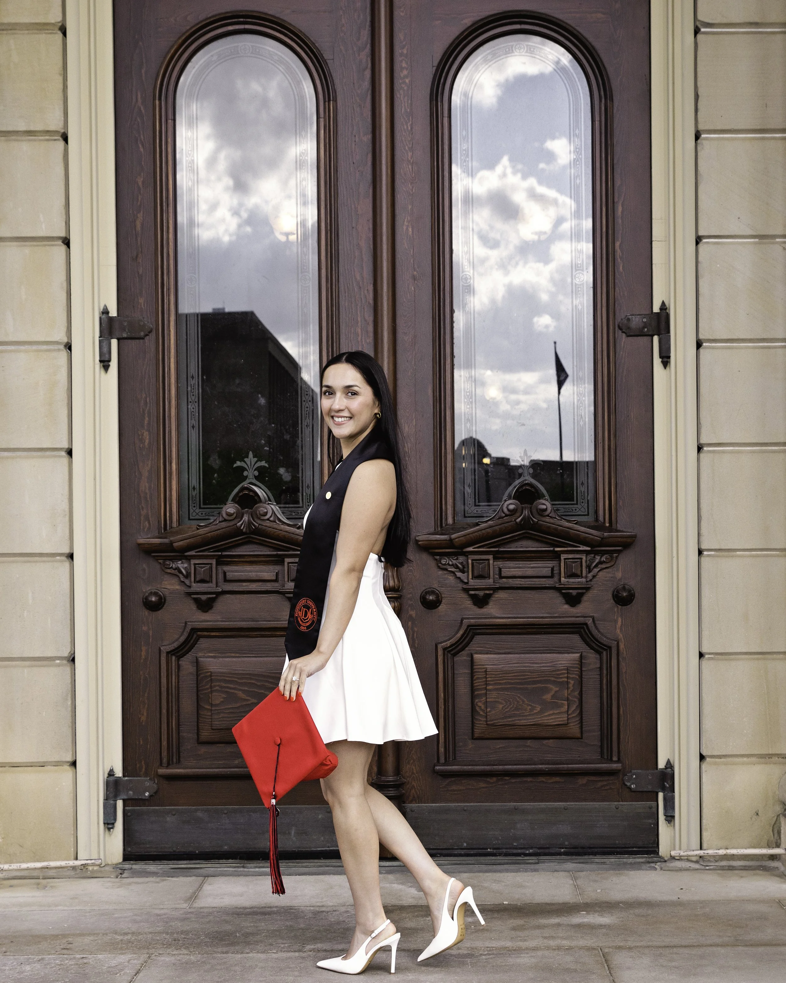 Young woman in a black top and white skirt standing outside in front of a large wooden door with glass panels, holding a red clutch, and wearing white high heels.