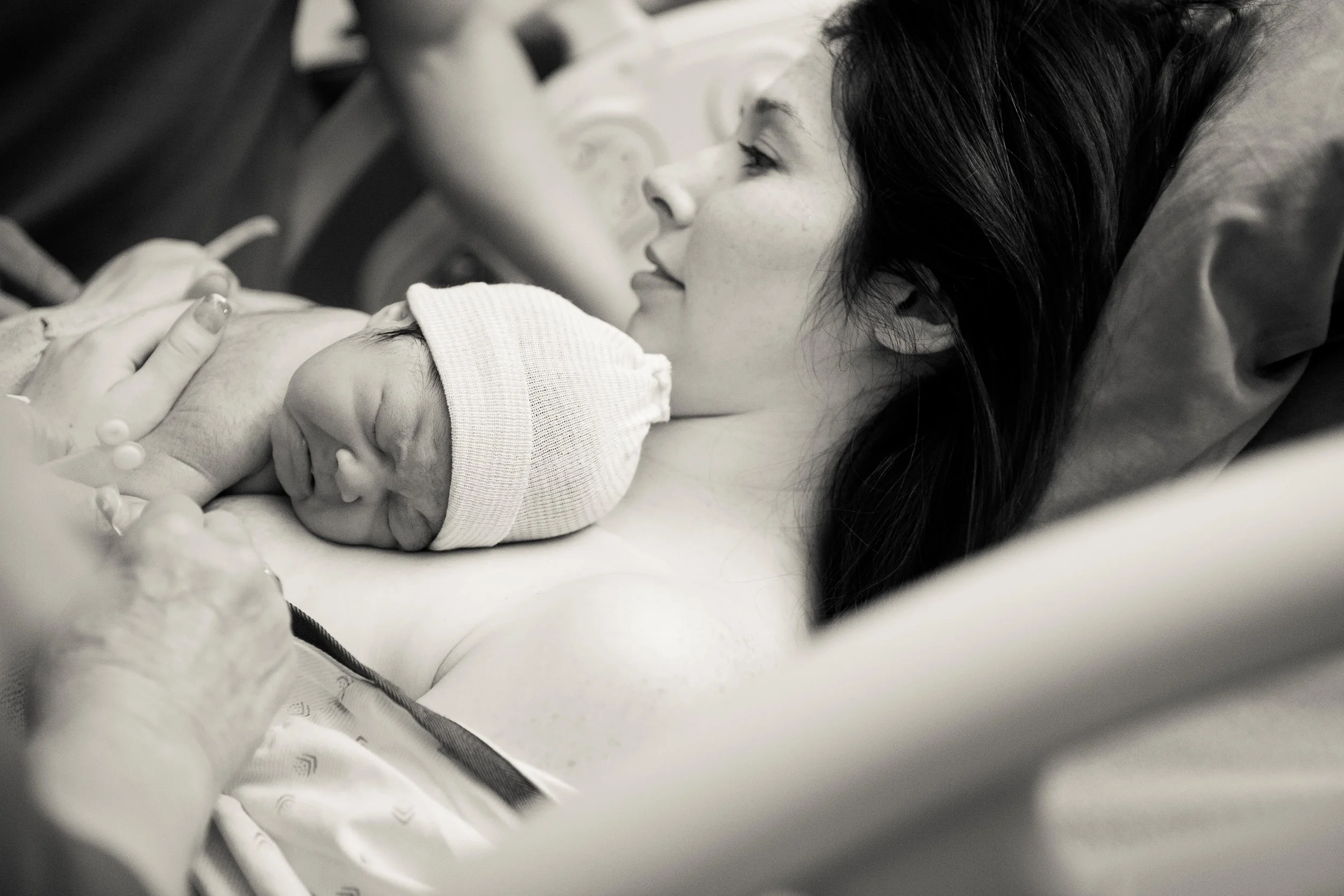 A woman lying in a hospital bed with her newborn baby, gently holding the baby's hand. The woman has dark hair and is gazing at the baby, who is wearing a hat.