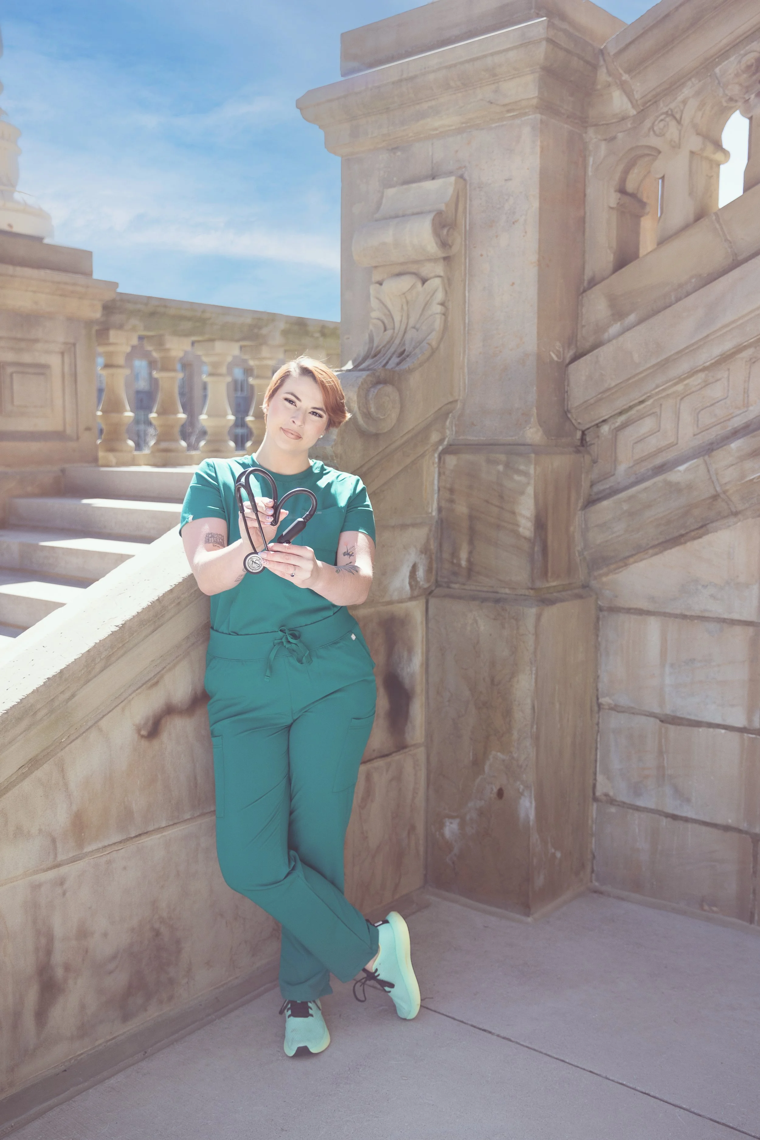A woman with short, reddish-brown hair, wearing medical scrubs, stands outdoors against a stone staircase with ornate details. She is holding a stethoscope and has a tattoo on her left arm. The sky is blue with some clouds.