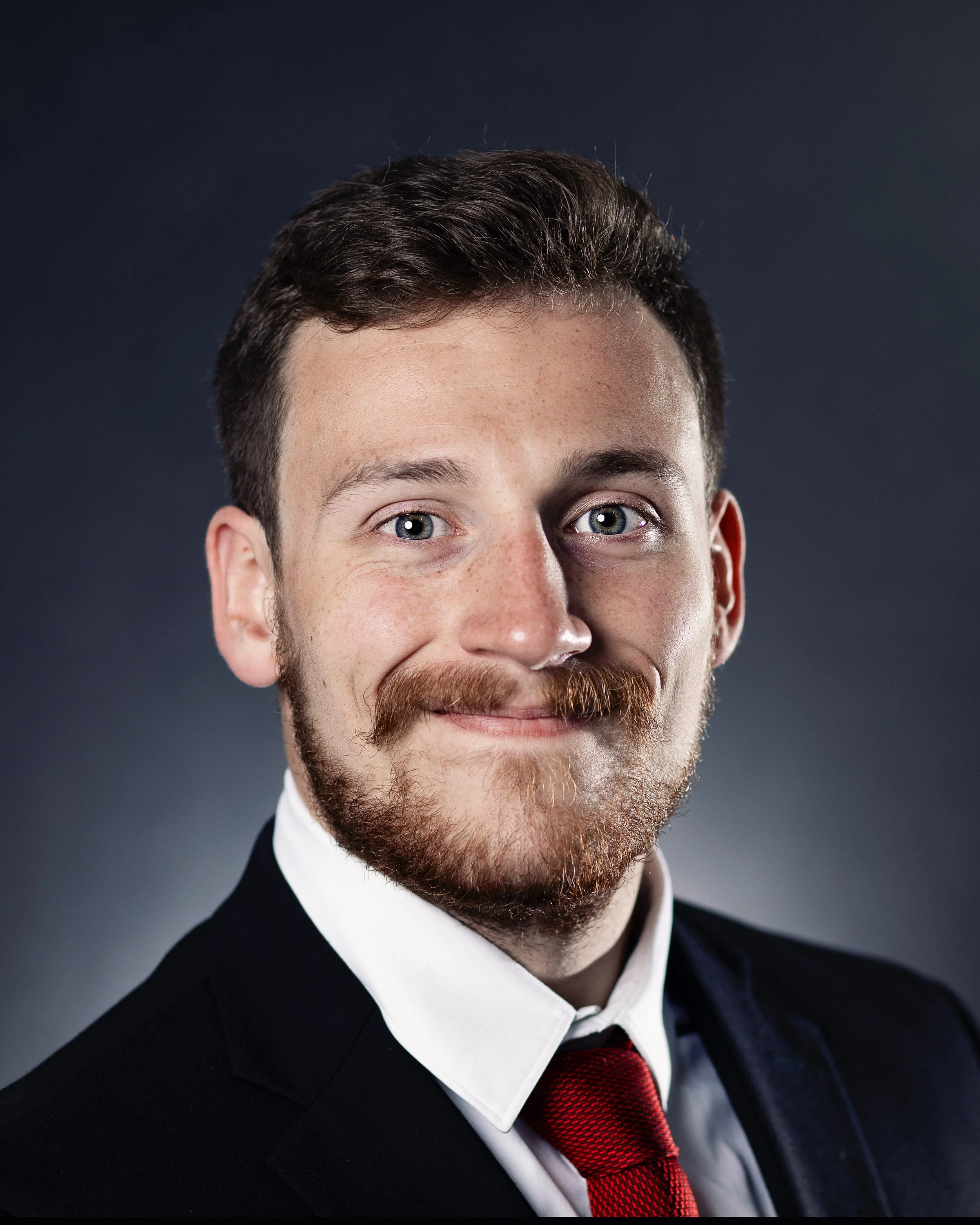 Close-up portrait of a man with short dark hair, gray eyes, a full beard, wearing a black suit, white shirt, and red tie, against a dark background.