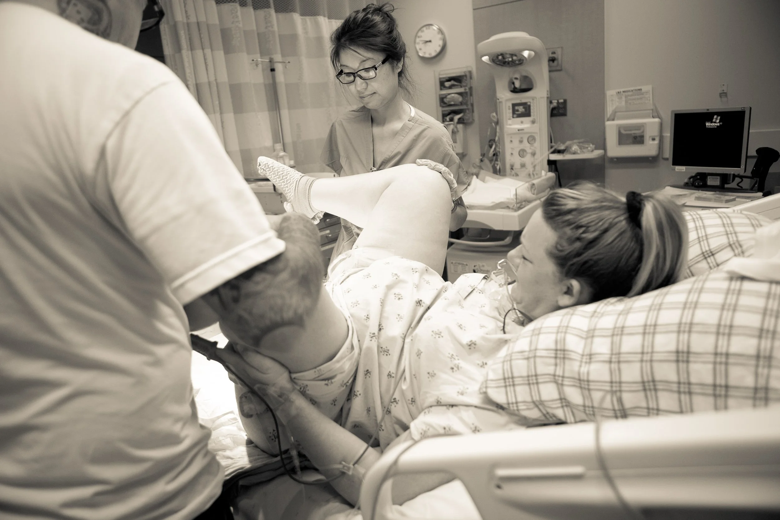 A medical professional, wearing glasses and scrubs, attends to a patient lying in a hospital bed. The patient, with an IV in her arm, is holding the doctor’s hand. Hospital medical equipment and monitors are visible in the background.