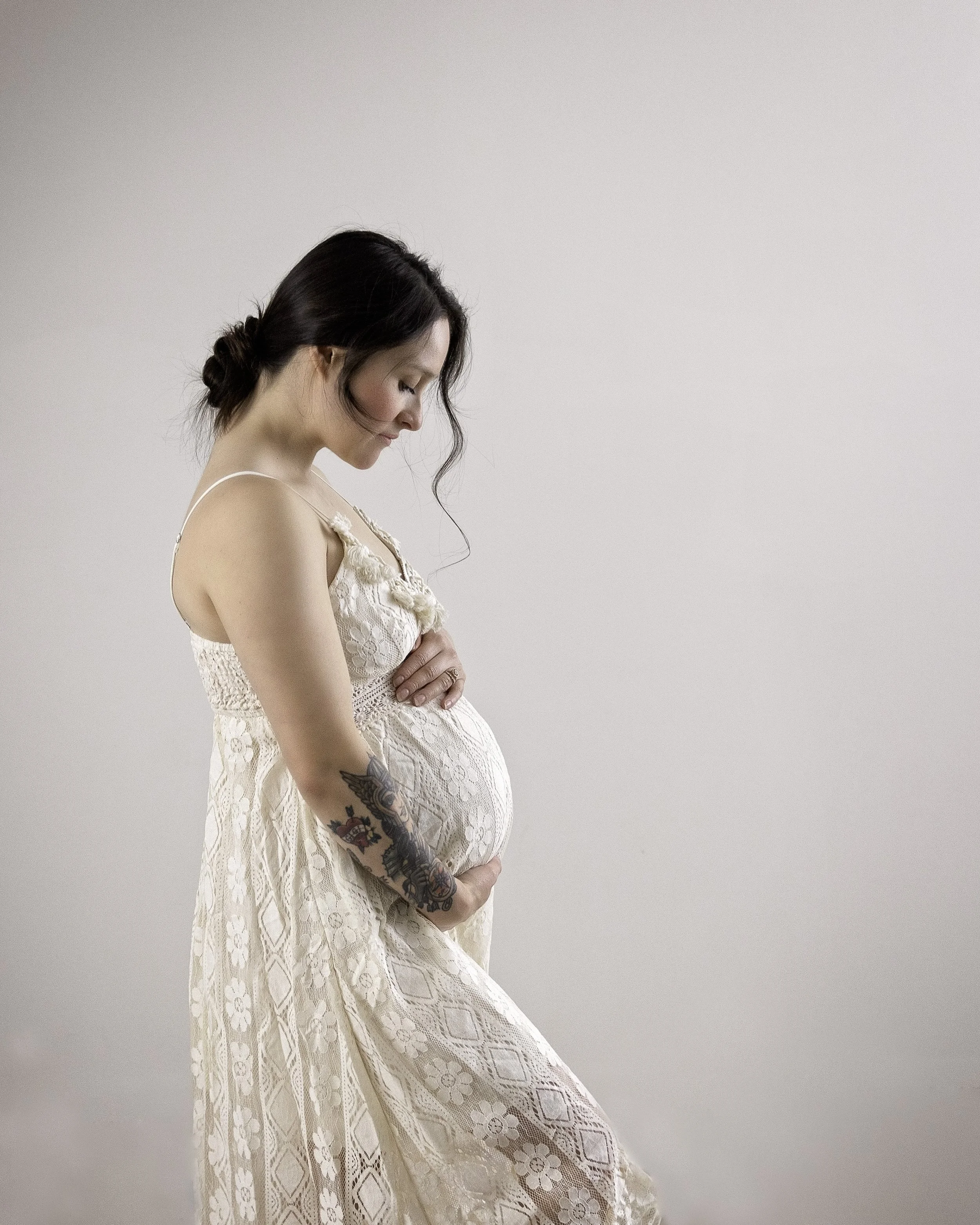 A pregnant woman with dark hair styled in a loose bun, tattoos on her left arm, wearing a light-colored lace dress, standing in profile with her head bowed and hands resting on her belly against a plain light background.