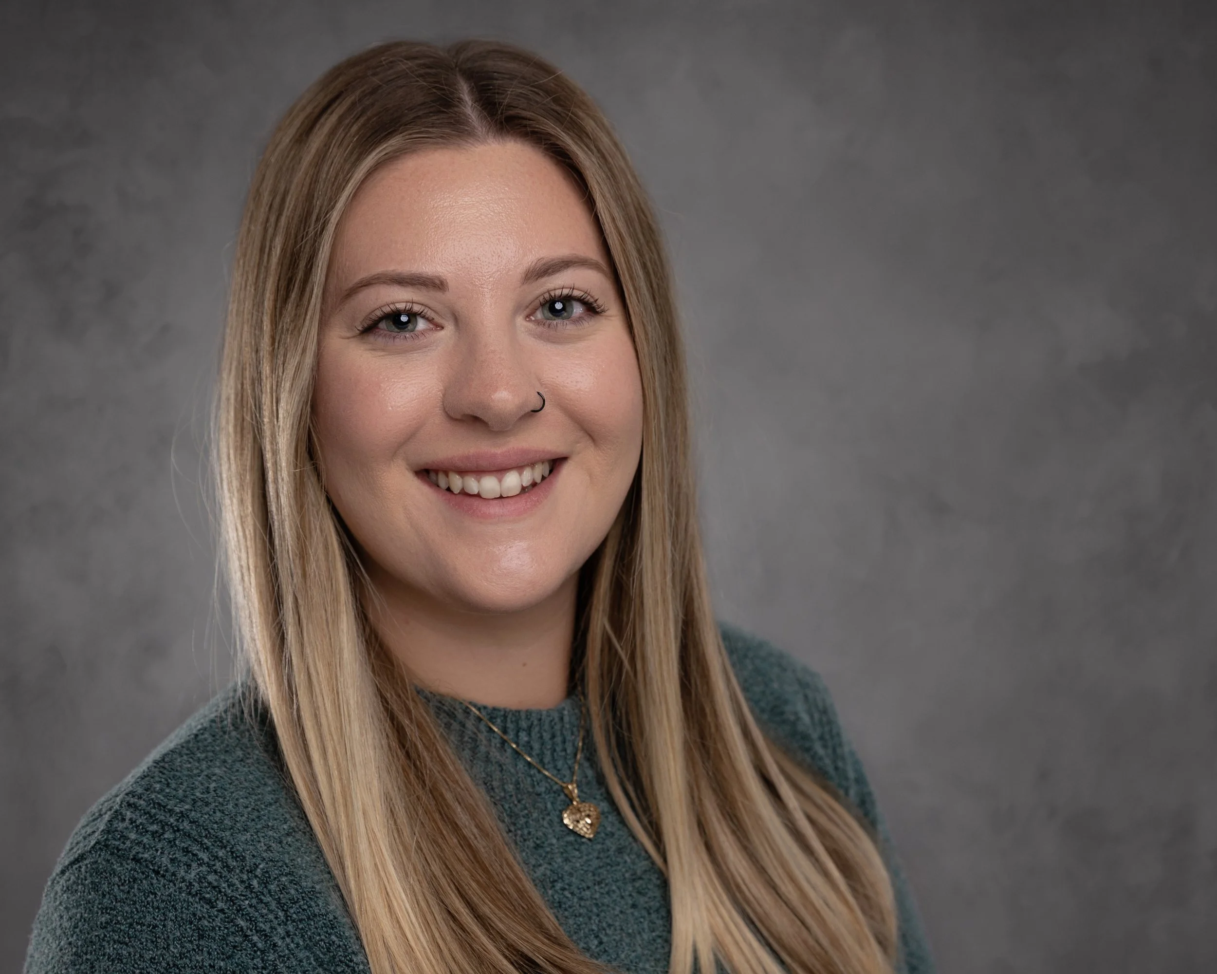 A young woman with long blonde hair, blue eyes, and a nose piercing smiles at the camera. She is wearing a dark green sweater and a gold necklace with a heart-shaped pendant. The background is a gray textured wall.