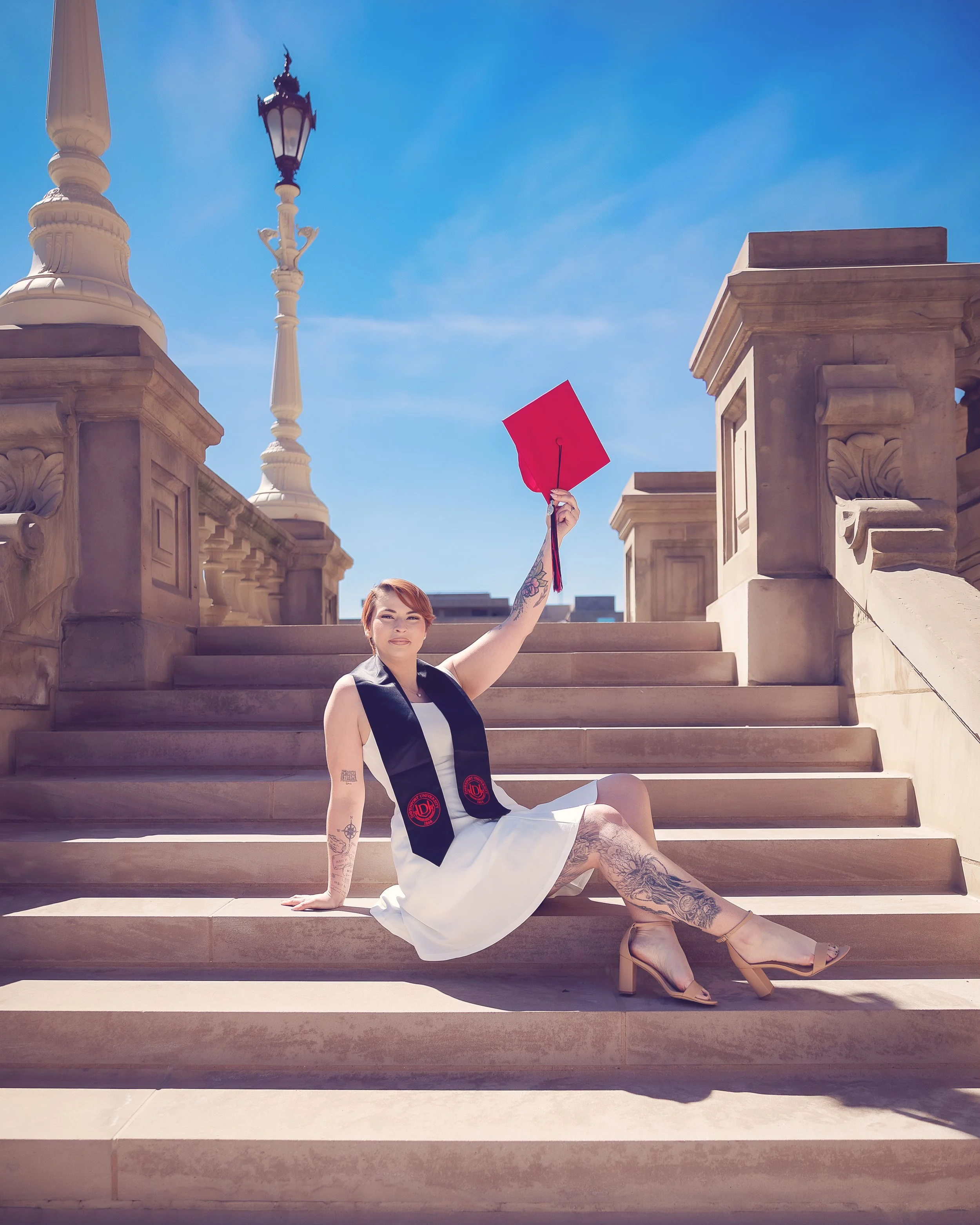 A woman with tattoos on her arm and leg sitting on outdoor steps, holding a red graduation cap in the air. She is wearing a sleeveless black and white dress and nude high heels. The background features stone balustrades and a blue sky.