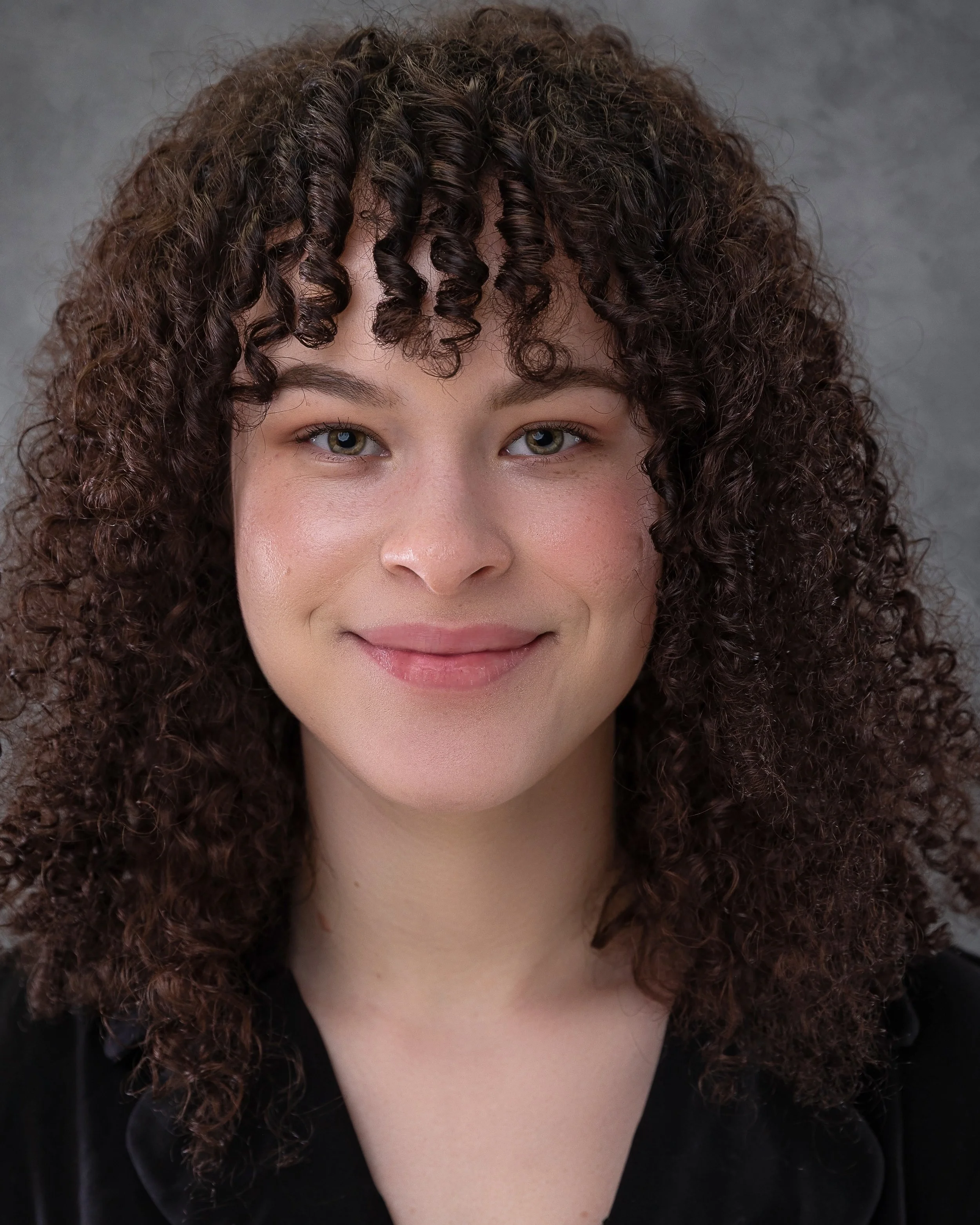 Close-up of a woman with curly dark brown hair, fair skin, and light-colored eyes, wearing a black top, against a gray background.