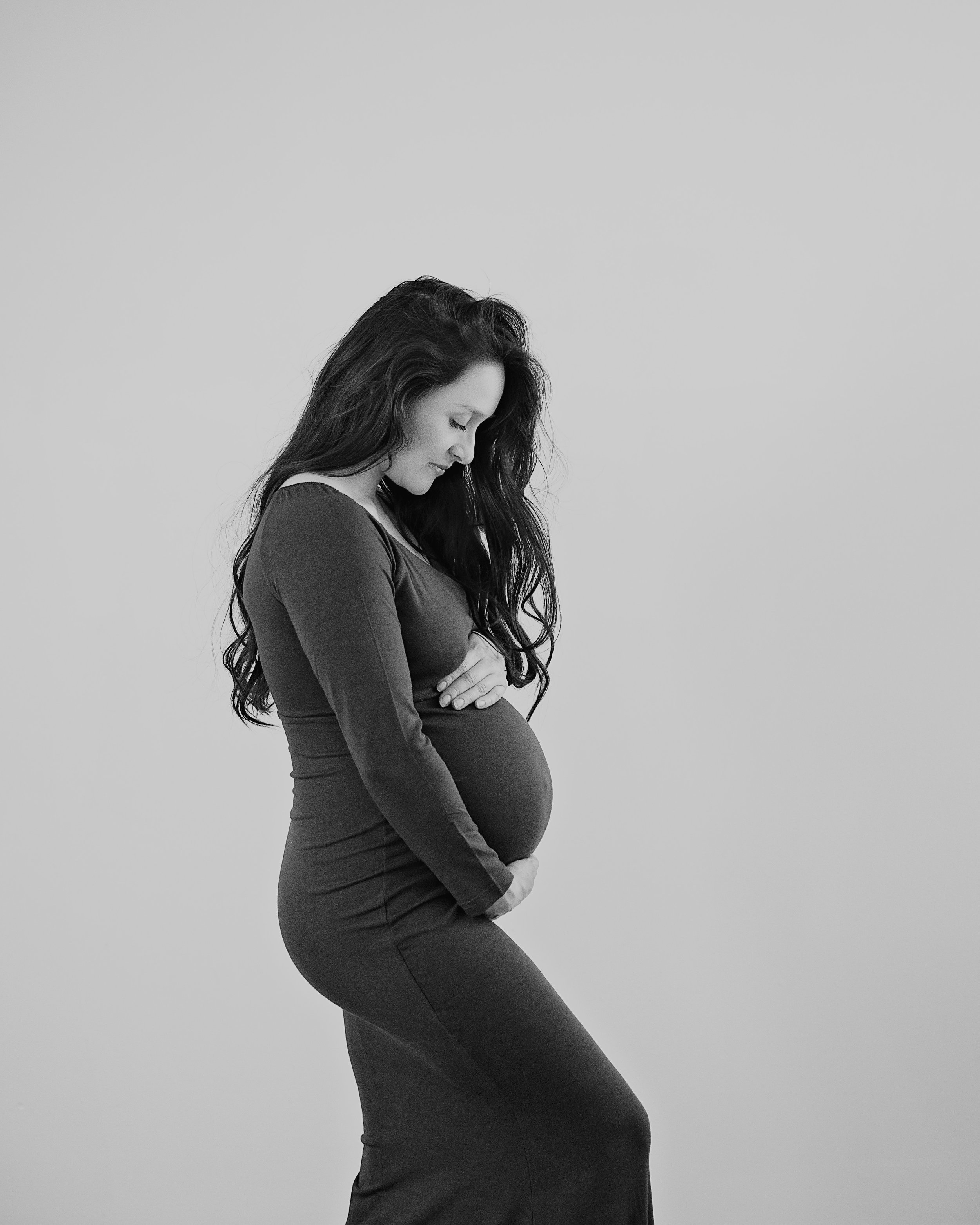 A pregnant woman in a form-fitting dress with long, dark, wavy hair, standing against a plain, light background, looking down with her arms crossed over her belly.