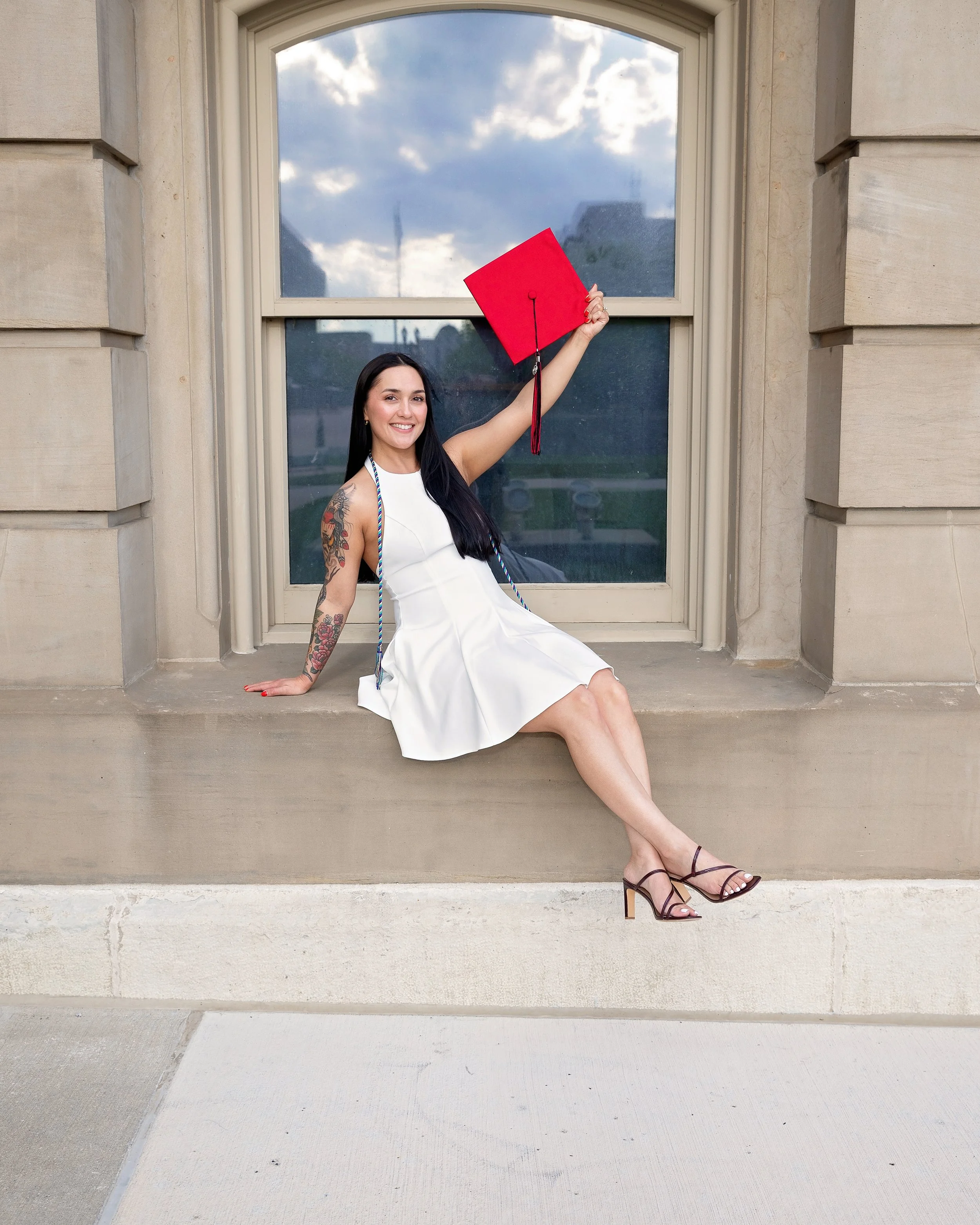A woman sitting on a stone ledge in front of a window with an arched top, holding a red graduation cap up in the air, wearing a white dress and high heel sandals, smiling at the camera, with tattoos on her left arm.