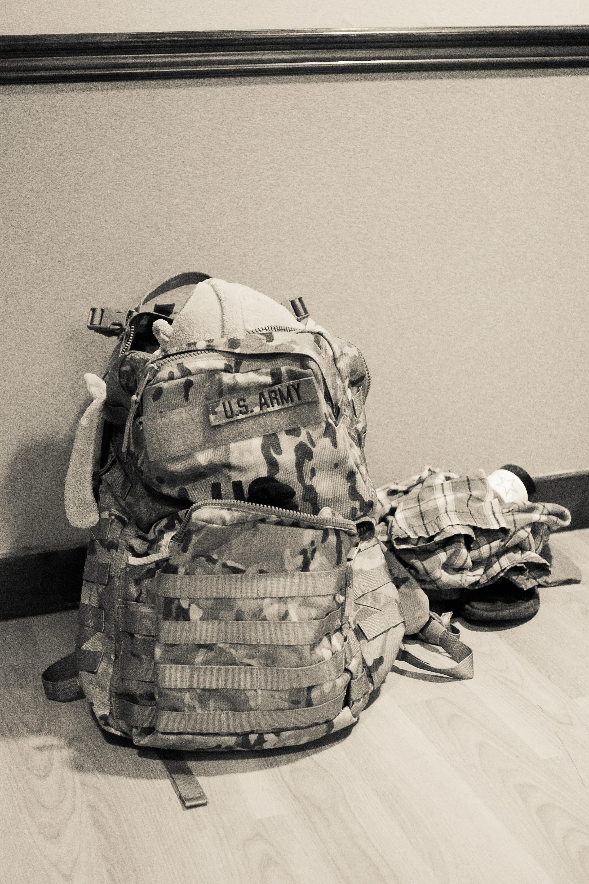 A U.S. Army camouflage backpack placed on the floor with a stuffed bear and a baseball cap on top, and various items scattered nearby, against a plain wall.