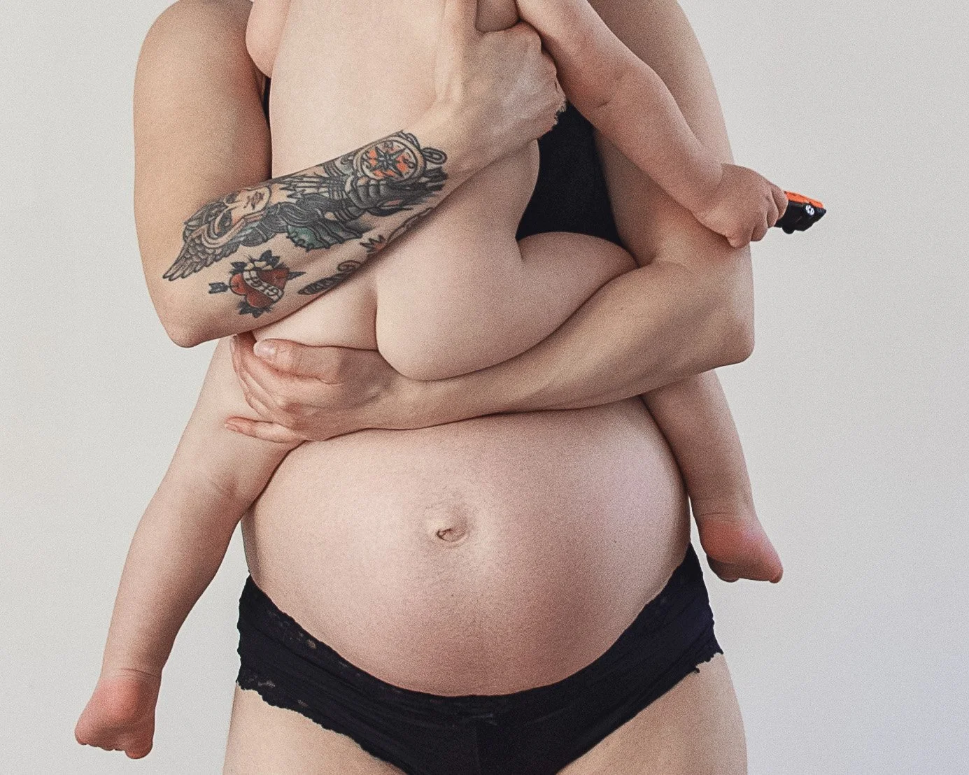 A person holding a child, with the person in black underwear and a tattoo on their arm. The child is holding a toy.