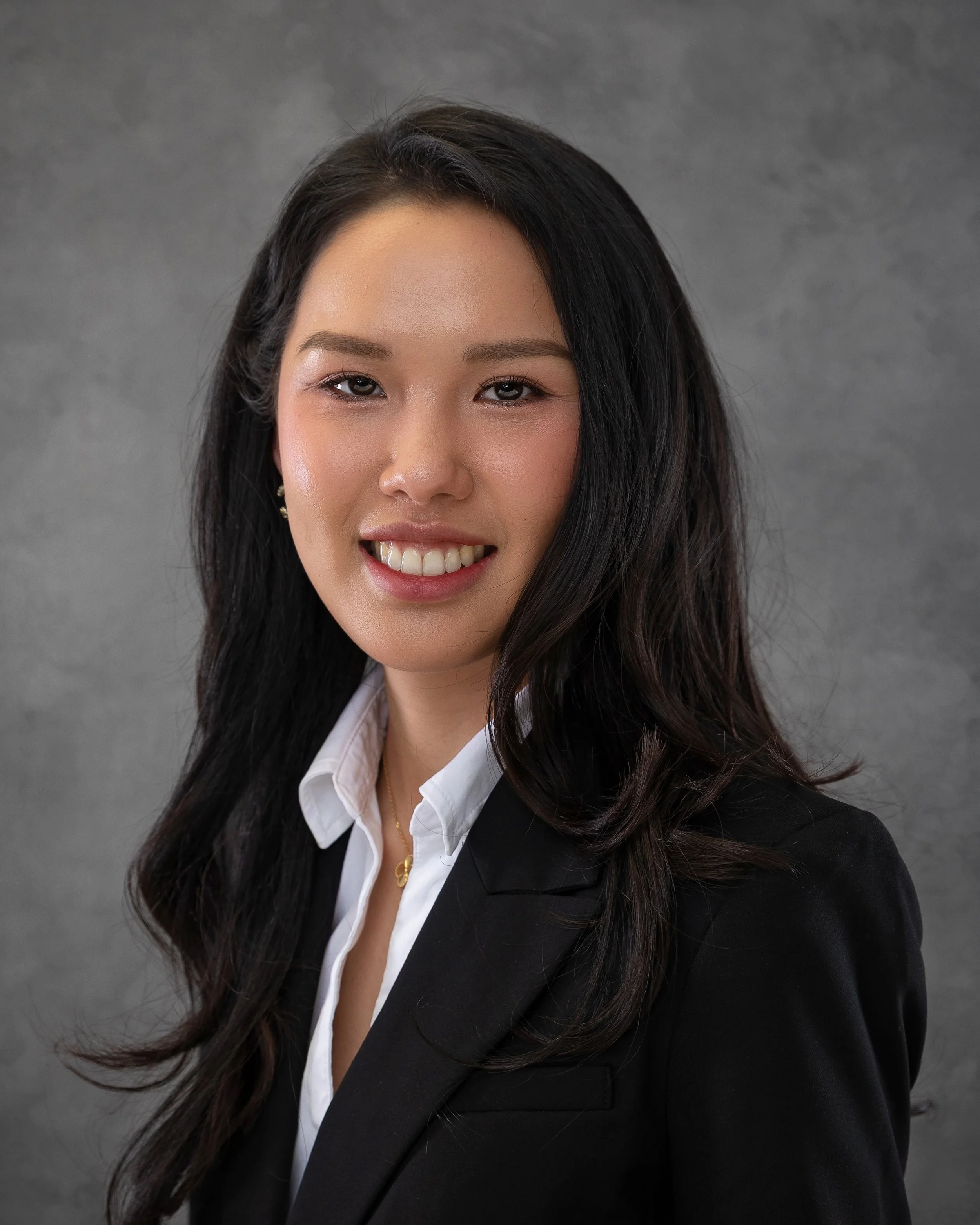A woman with long black hair smiling, wearing a black blazer over a white shirt, set against a gray background.