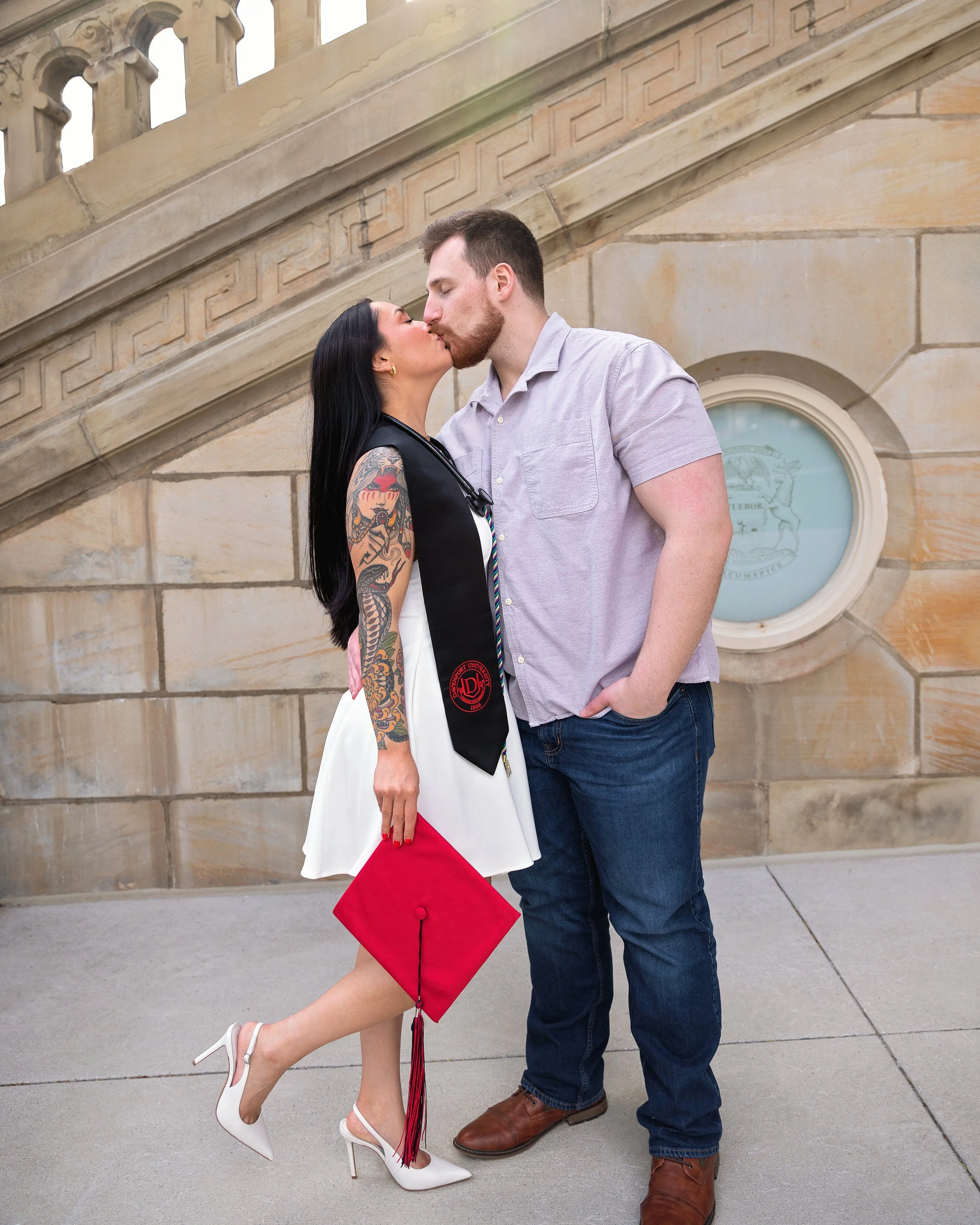 A woman and a man kissing outdoors, the woman wearing a white dress, a black graduation stole, and high-heeled white shoes, holding a red diploma cover, with her leg slightly lifted. The man is dressed in a light-colored collared shirt, dark jeans, a