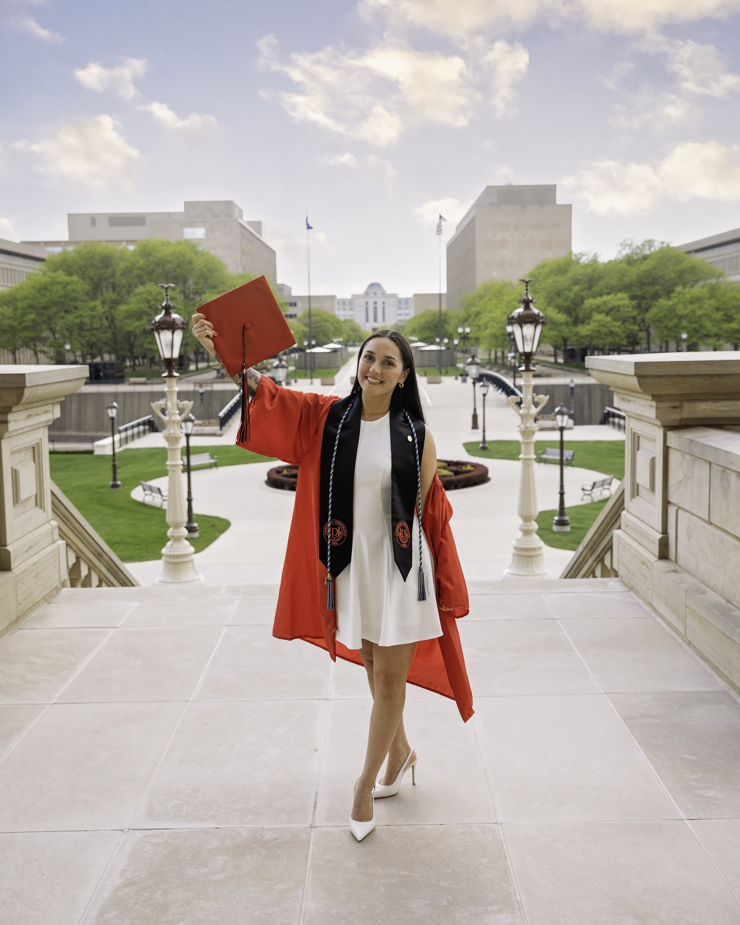 A young woman in a white dress and high heels wearing a red graduation gown and cap, holding her cap in the air, standing on steps with a university campus in the background.