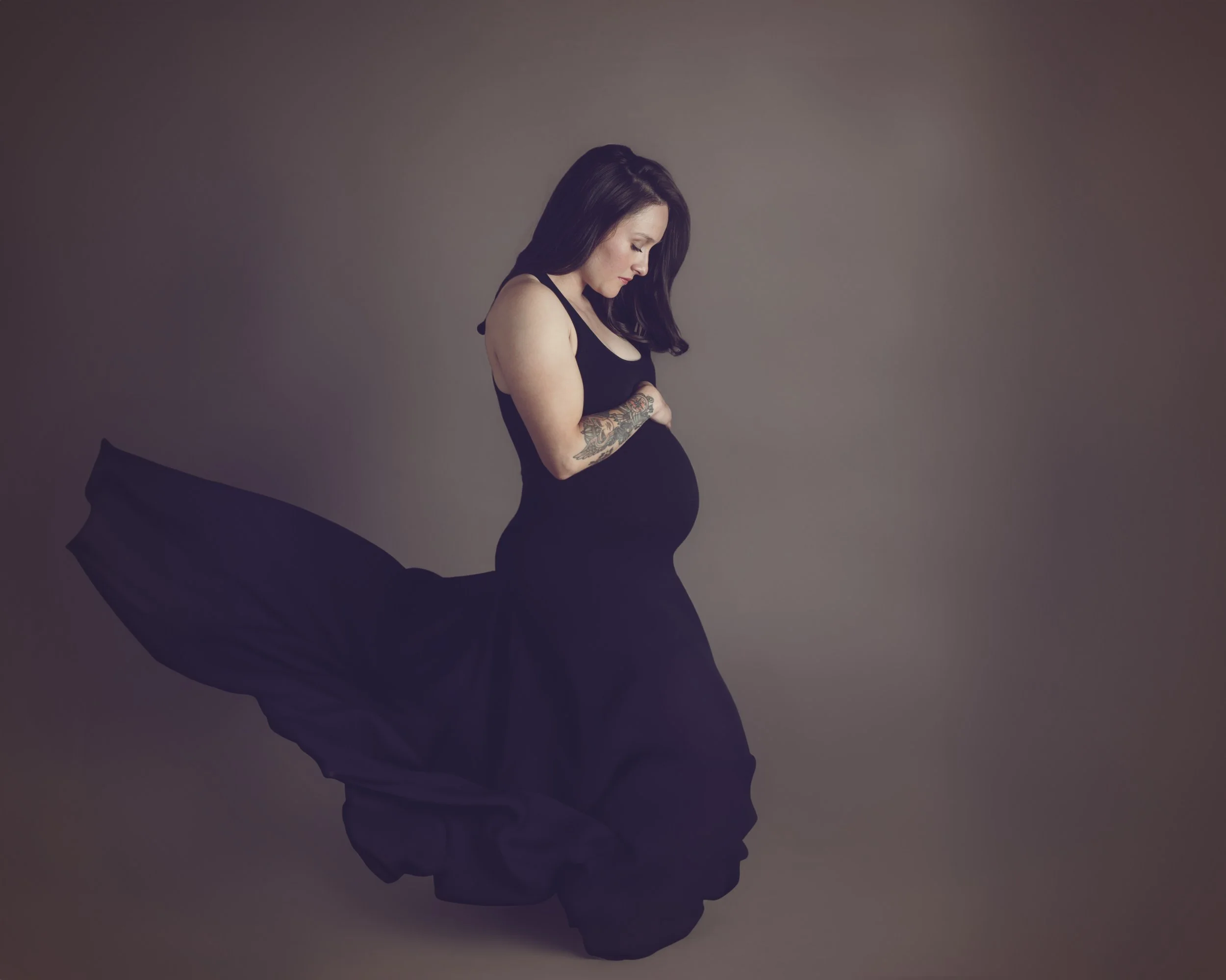 A woman with long dark hair and tattoos on her arm, wearing a black sleeveless dress, standing in profile against a plain background.