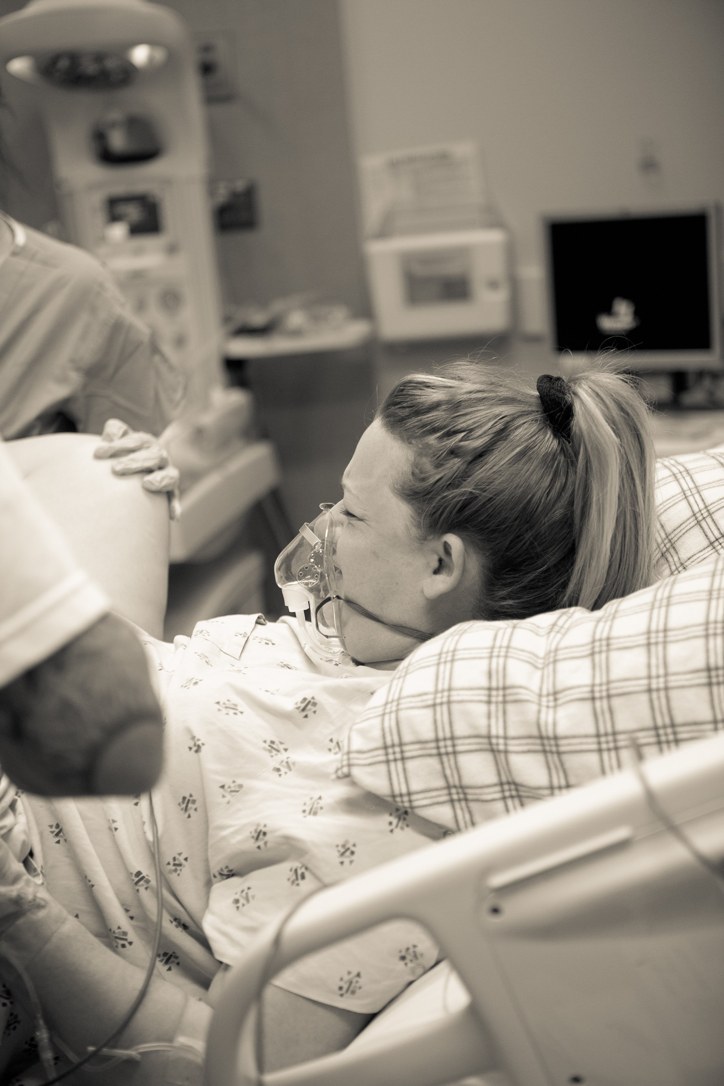 A woman with a ponytail lying in a hospital bed, connected to a breathing mask, with medical equipment in the background.