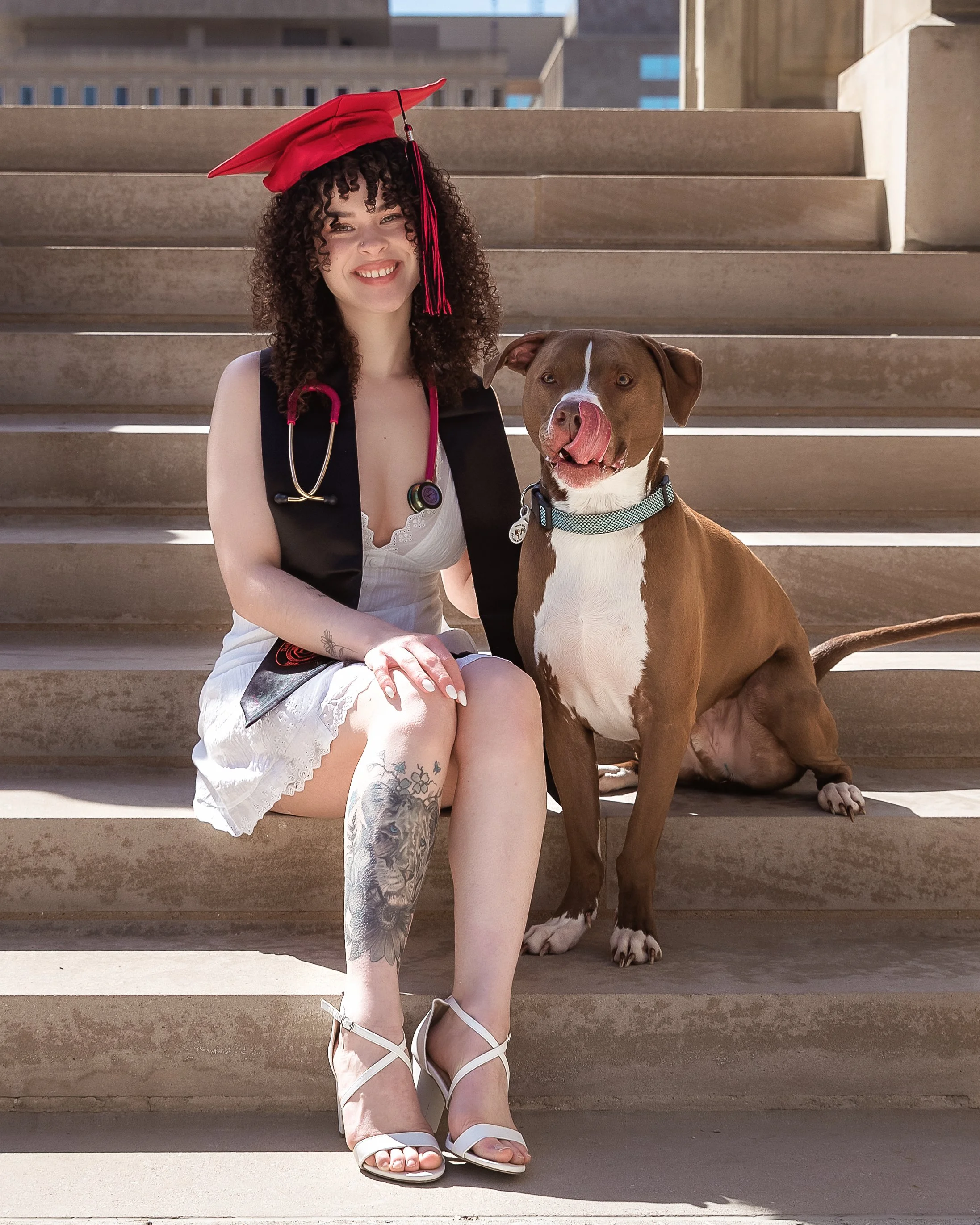 A woman with curly hair wearing a red graduation cap, a stethoscope, and a white lace dress sitting on outdoor steps with a brown and white dog licking her face.