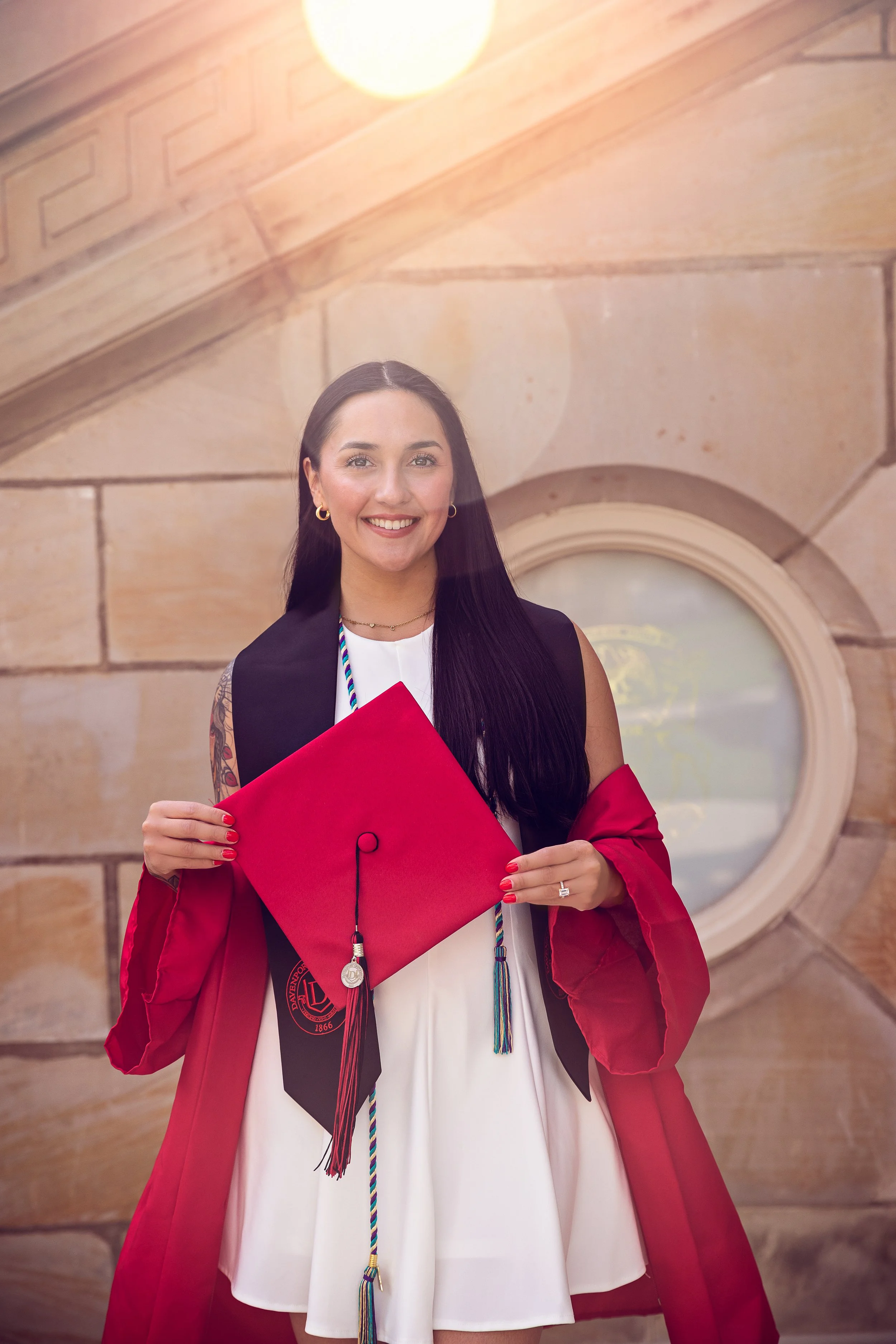A young woman dressed in a university graduation gown and holding a red diploma cover, standing in front of a stone building with a round window.
